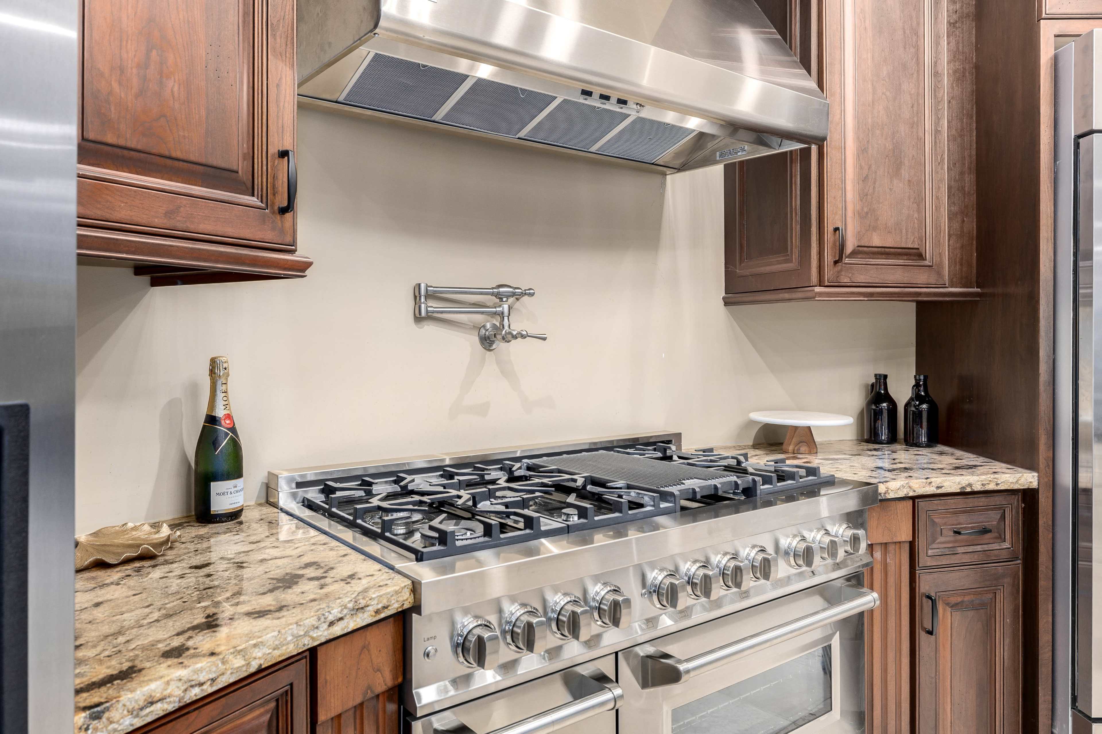 The image shows a modern kitchen featuring a large stainless steel gas range, a vent hood above it, and wooden cabinetry alongside granite countertops.