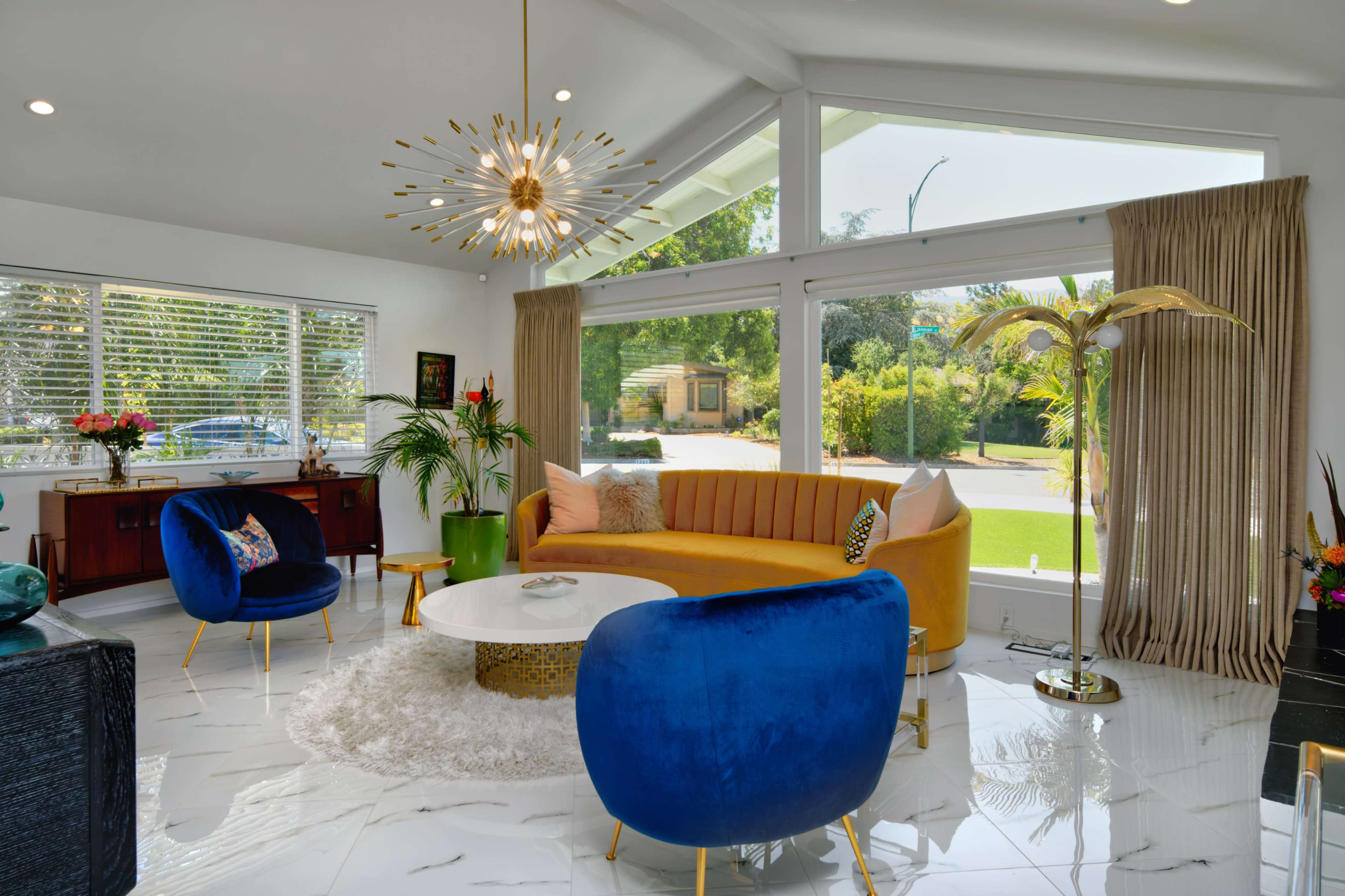 The image shows a modern living room featuring a yellow sectional sofa, blue accent chairs, a round coffee table, and large windows with views of the outdoors.
