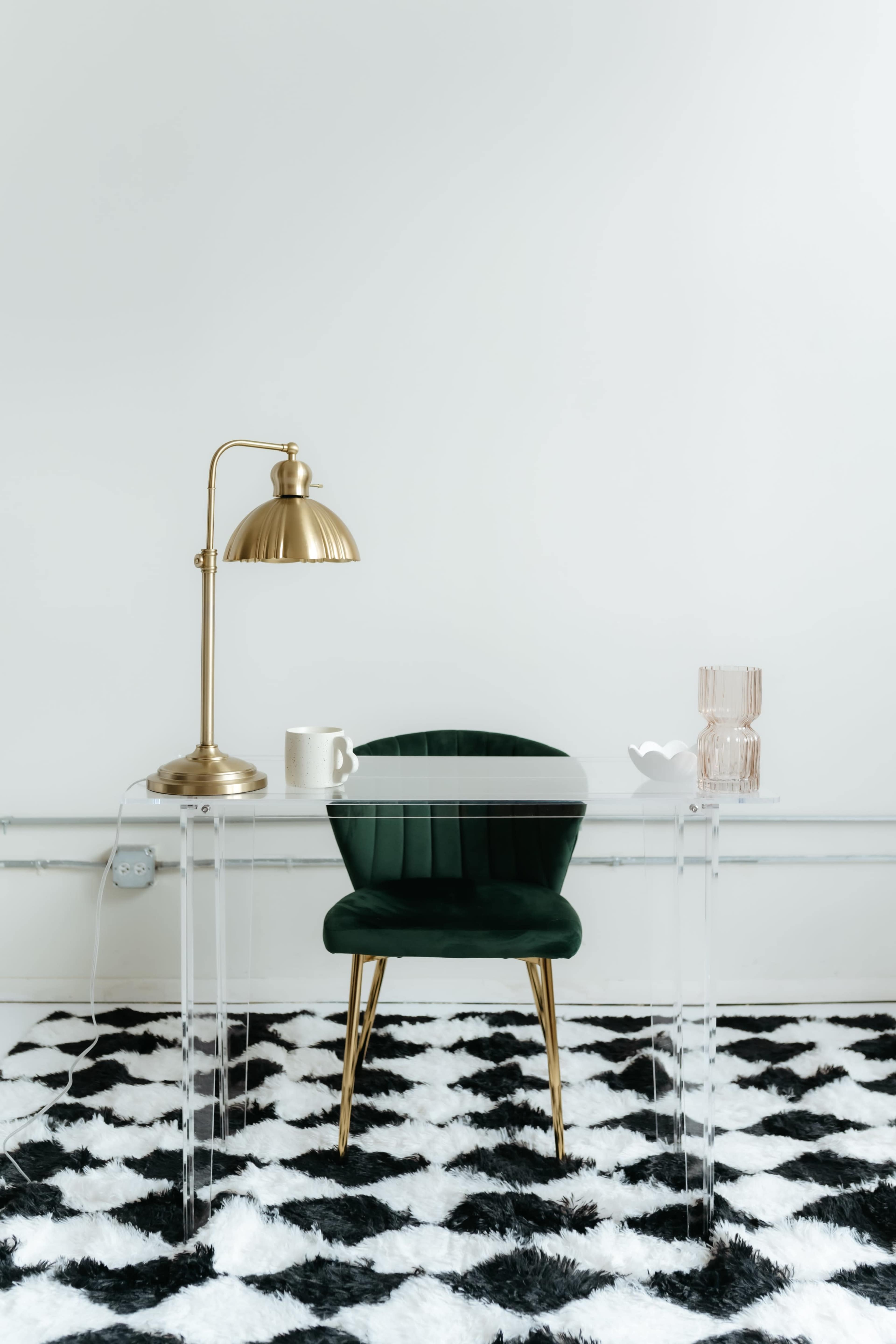 A clear glass desk with a green chair, a gold lamp, a white mug, and a decorative vase sits on a black and white striped rug.