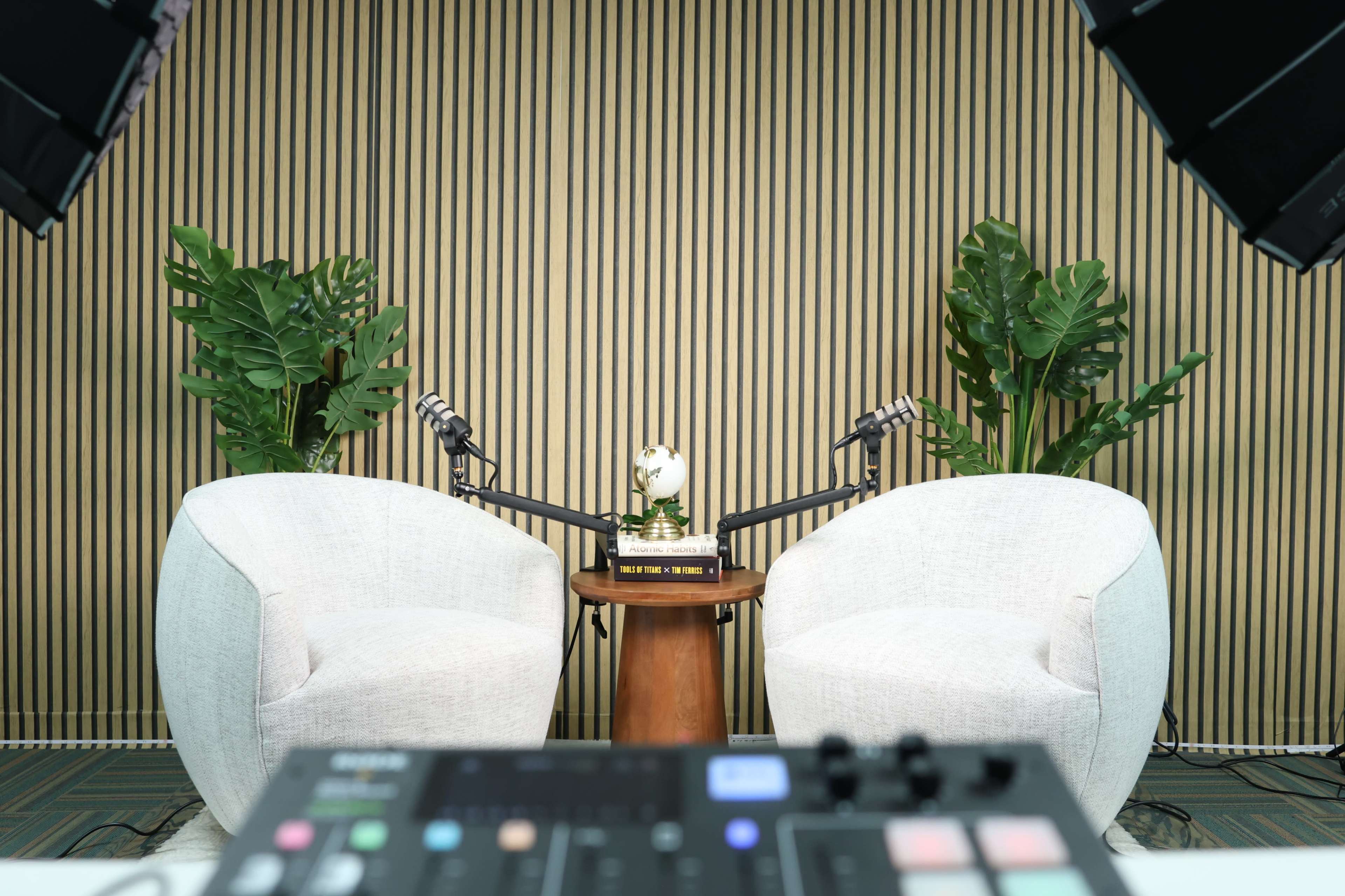 The image depicts a podcast studio setup with two white chairs, a small wooden table, a microphone setup, and green plants in the background against a striped wall.