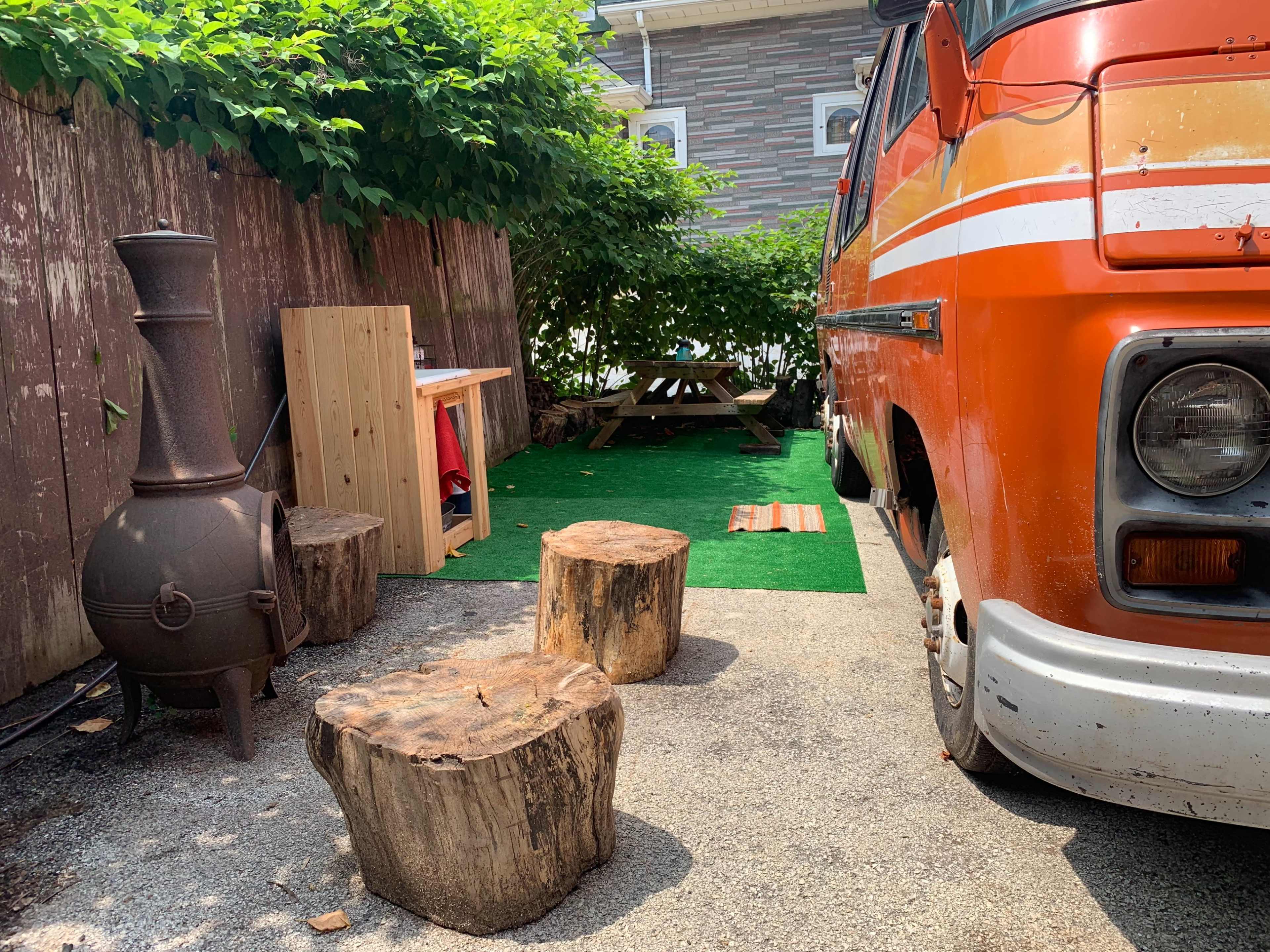 An orange van is parked next to a small outdoor seating area with wooden stools and a picnic table, surrounded by greenery and a stone path.