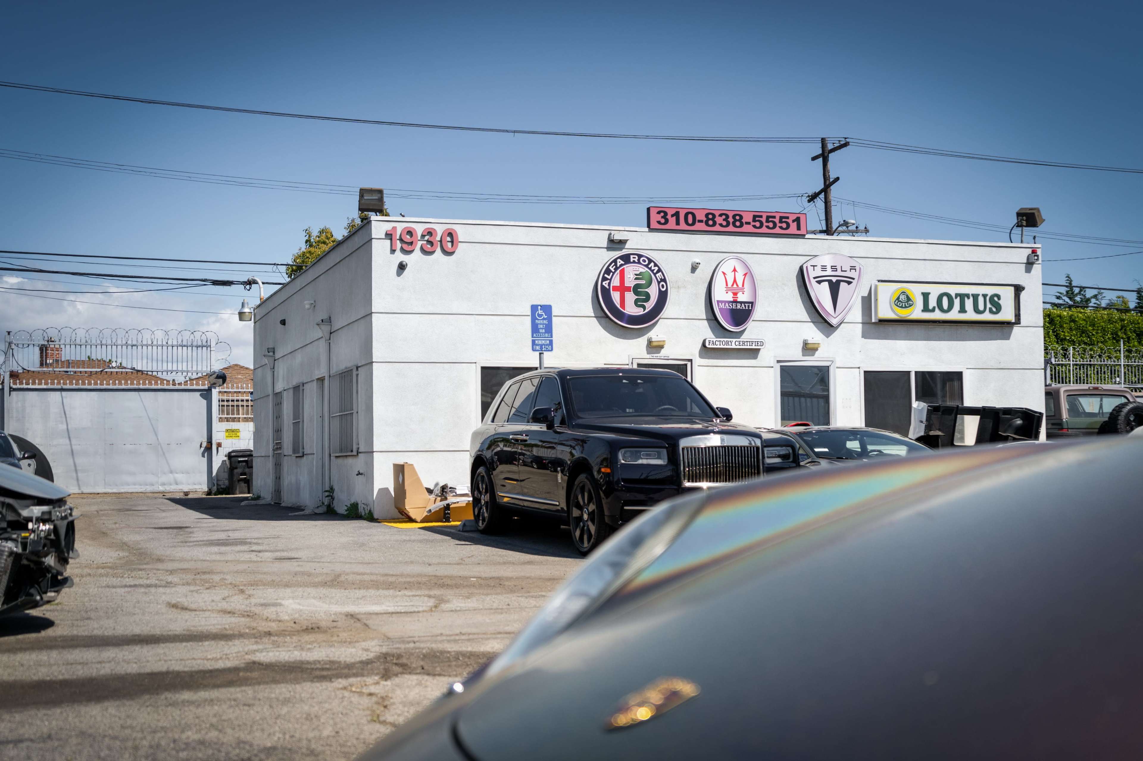 The image shows a car dealership with several luxury car brand logos displayed on the exterior wall of a white building, alongside a parked black Rolls-Royce.