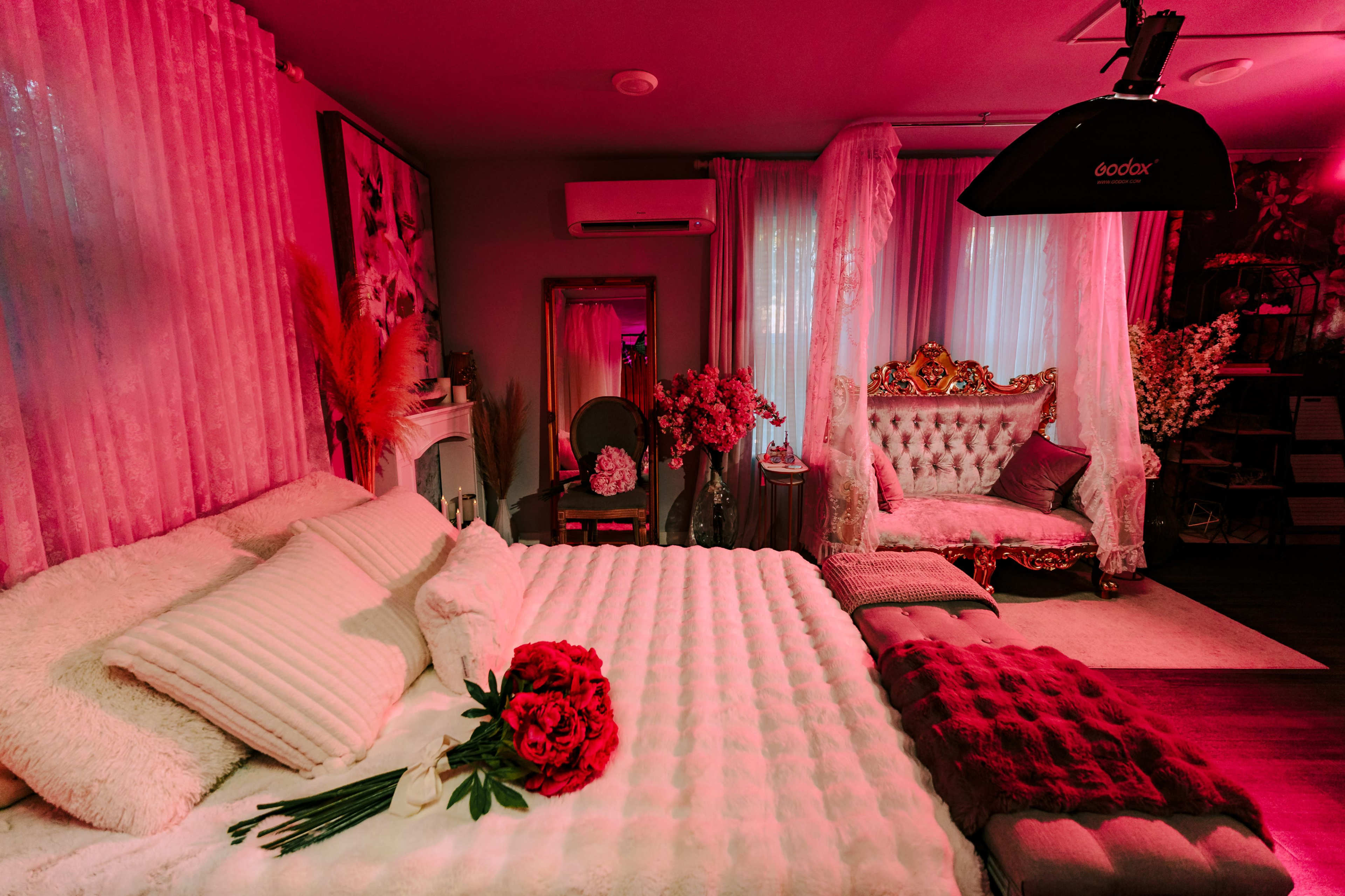 The image shows a styled bedroom featuring a plush bed with a white coverlet, a bouquet of red flowers on the bed, and an ornate seating area with decorative elements.