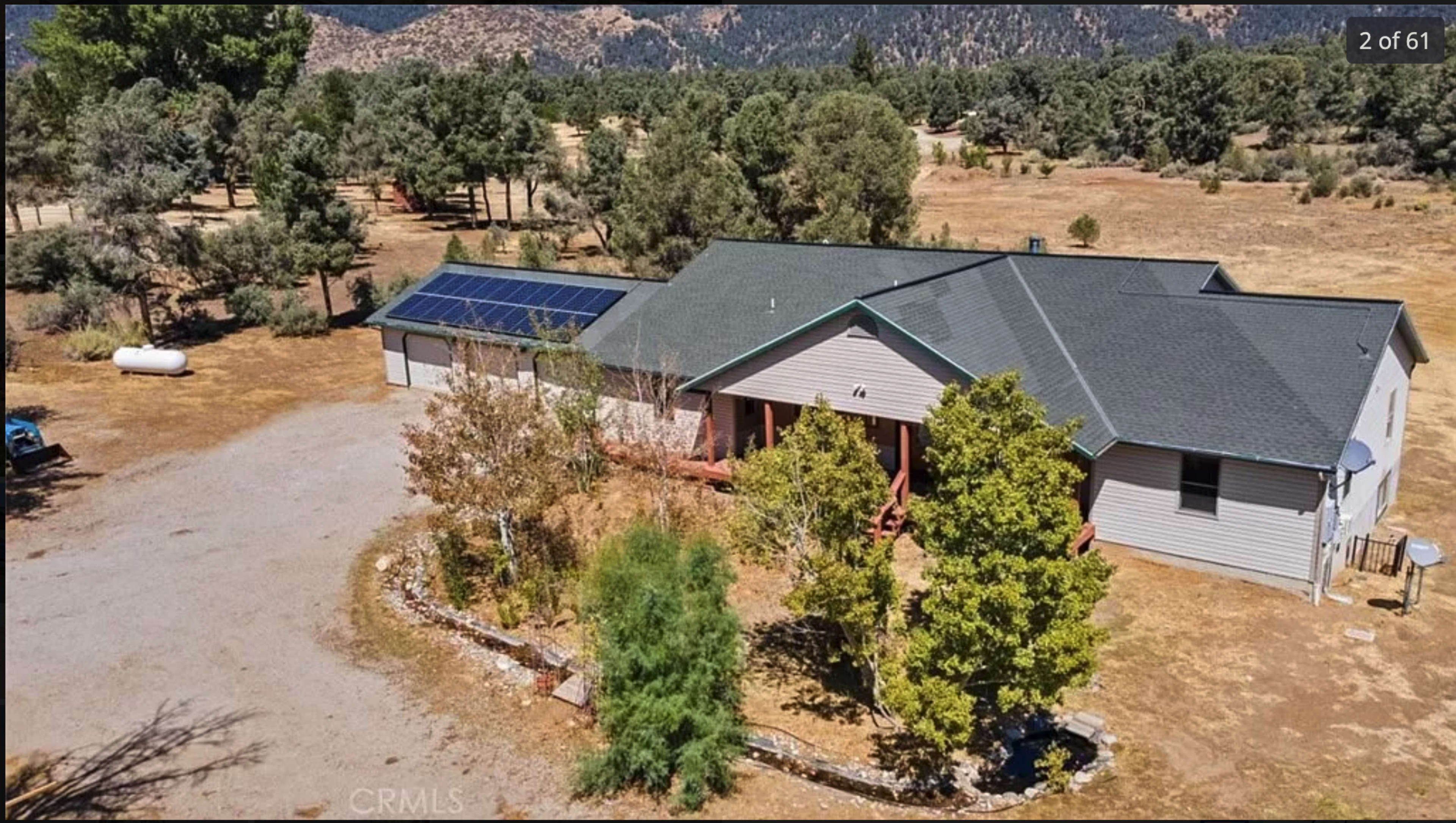 The image shows a single-story house with a solar panel on the roof, surrounded by trees and a large open area.