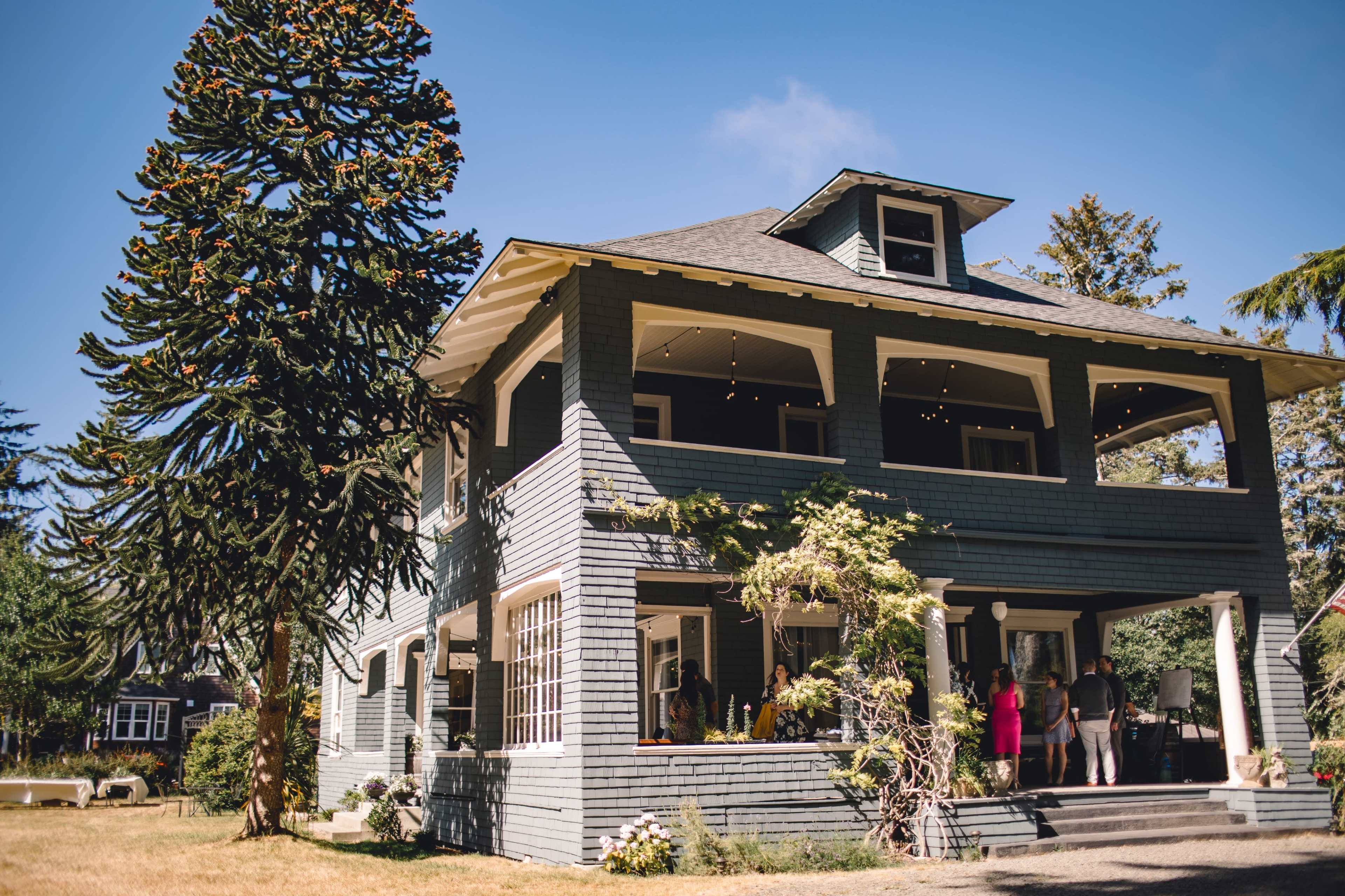 A large, two-story gray house with a sloped roof, large porch, and a tall tree in the foreground.