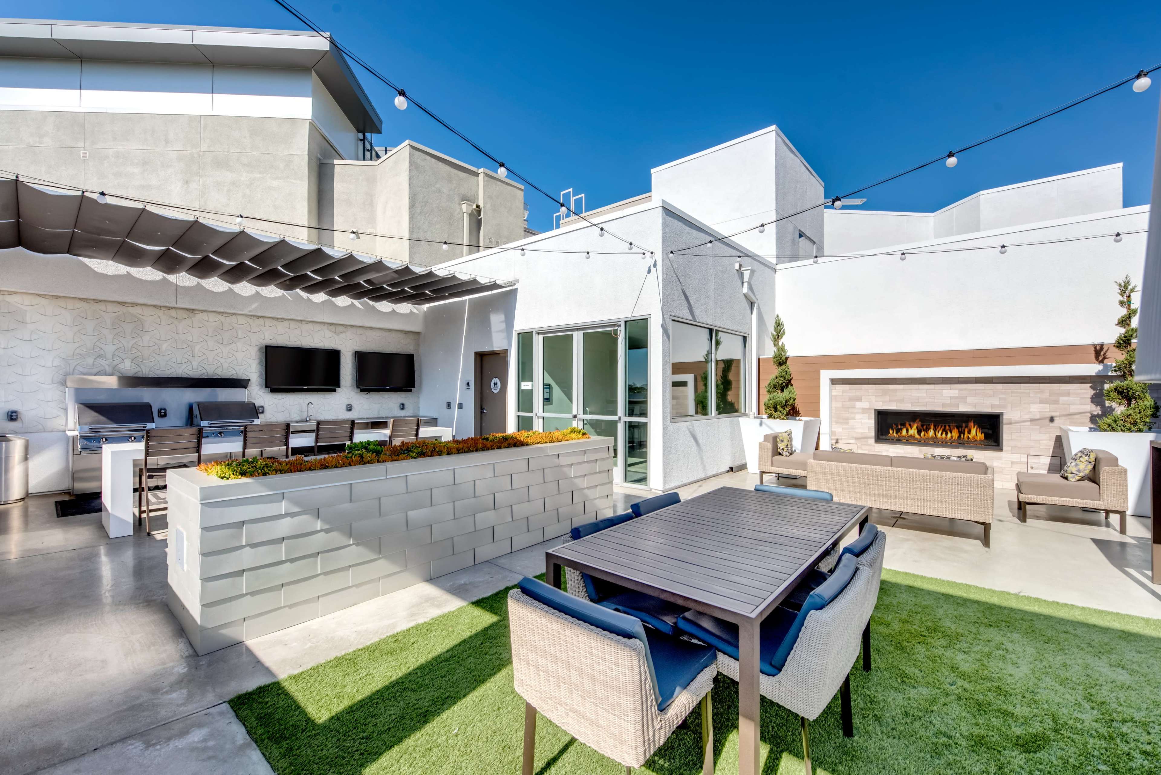 The image shows an outdoor patio area with a dining table, barbecue stations, and a fireplace surrounded by modern seating and greenery.