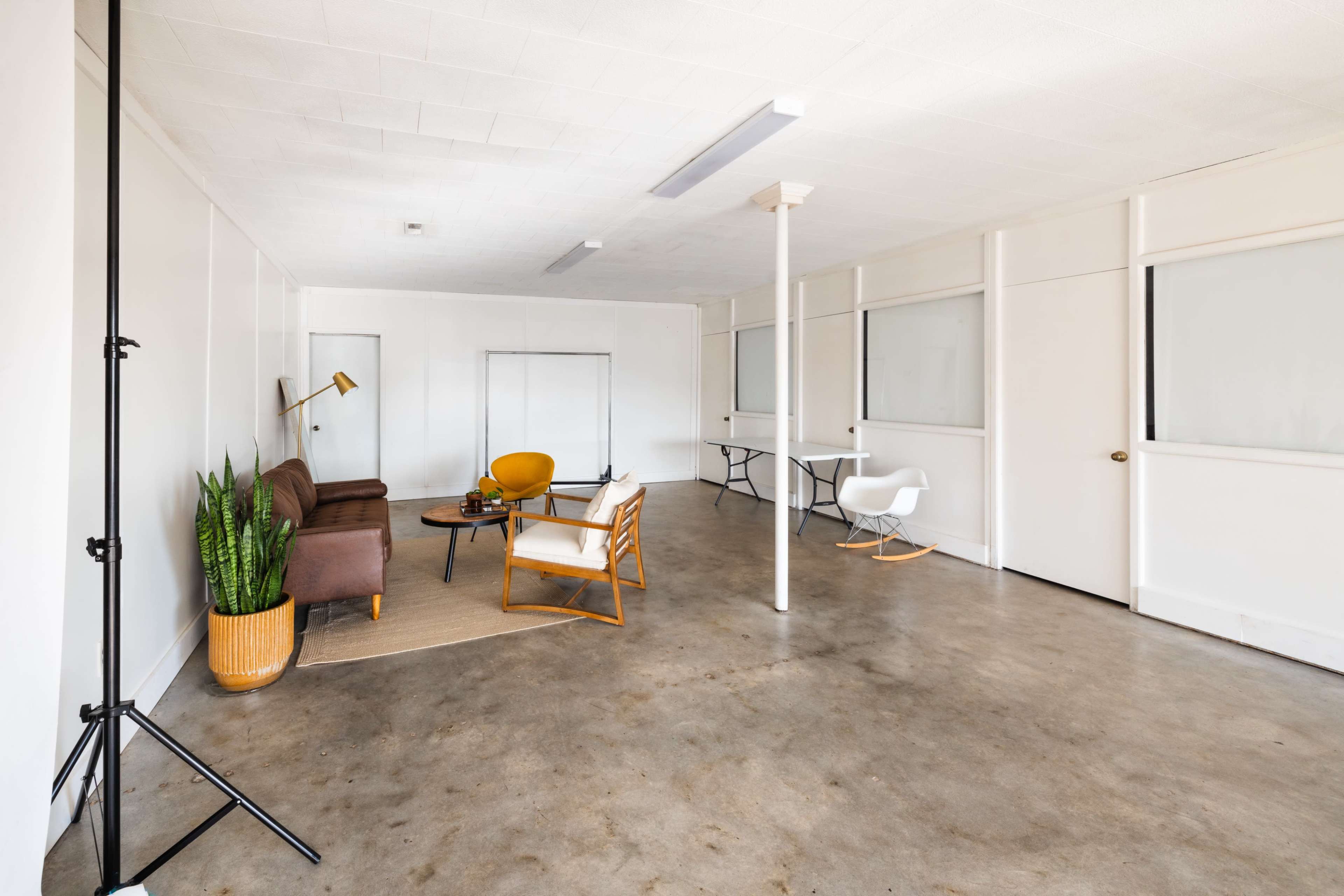 The image shows a spacious, uncluttered room with concrete flooring, featuring a small seating area with two chairs, a table, and a potted plant.