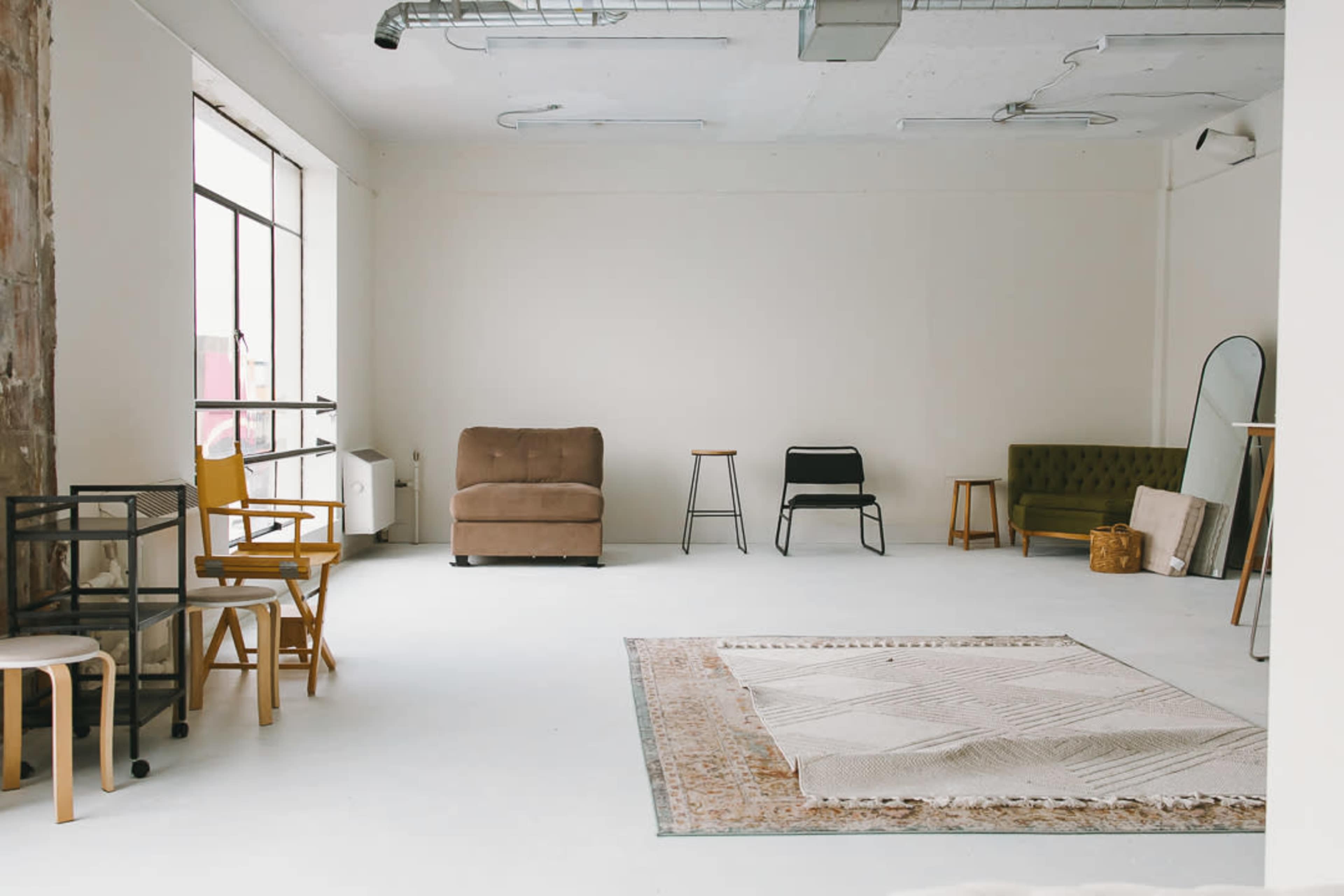 The image shows a spacious, minimalistic room with a light-colored floor, several pieces of furniture including chairs, a sofa, and a rug, and large windows allowing natural light to enter.