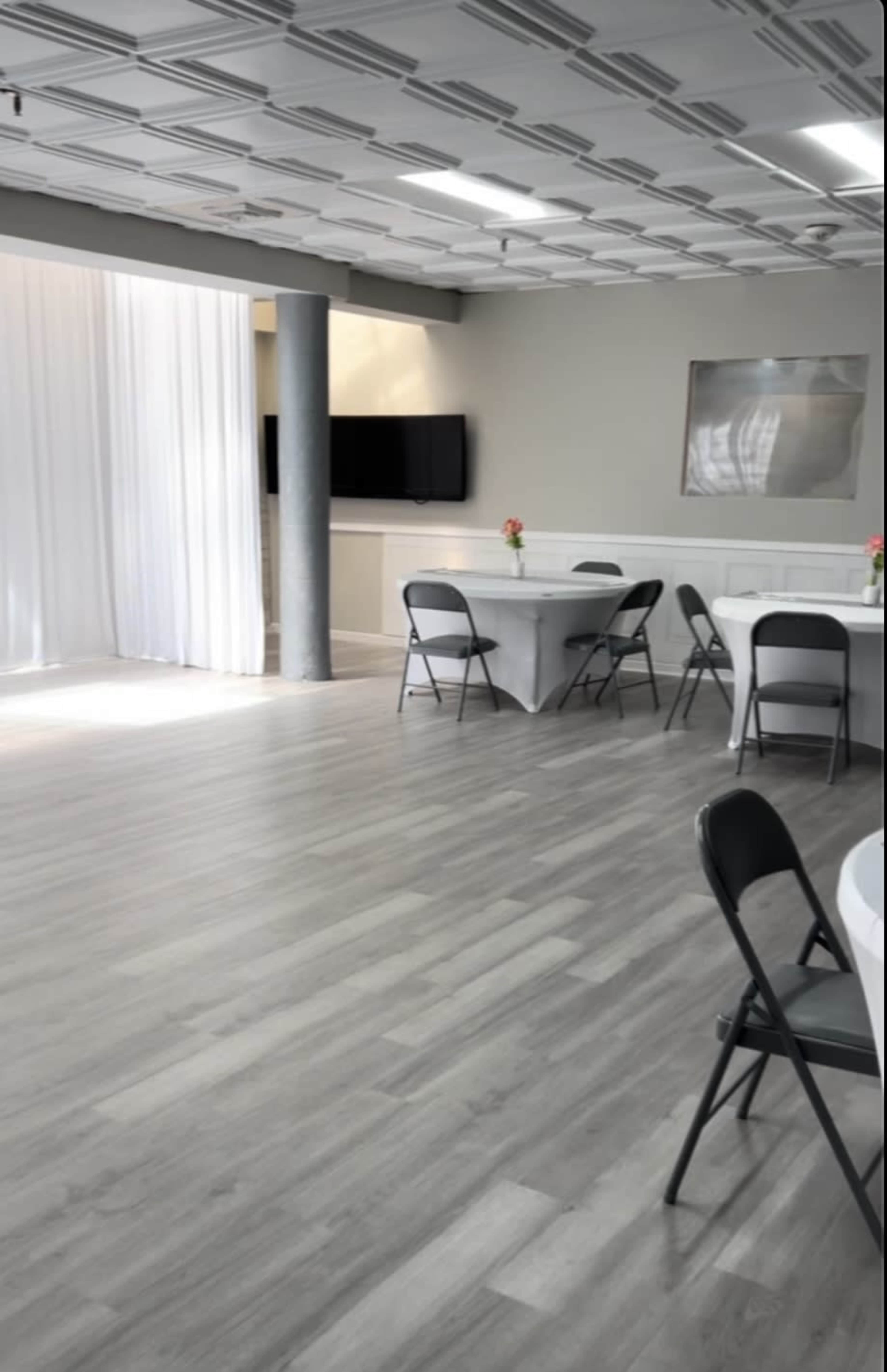 The image shows a spacious, well-lit indoor venue with gray laminate flooring, several round tables covered in white tablecloths, and black folding chairs arranged around them.