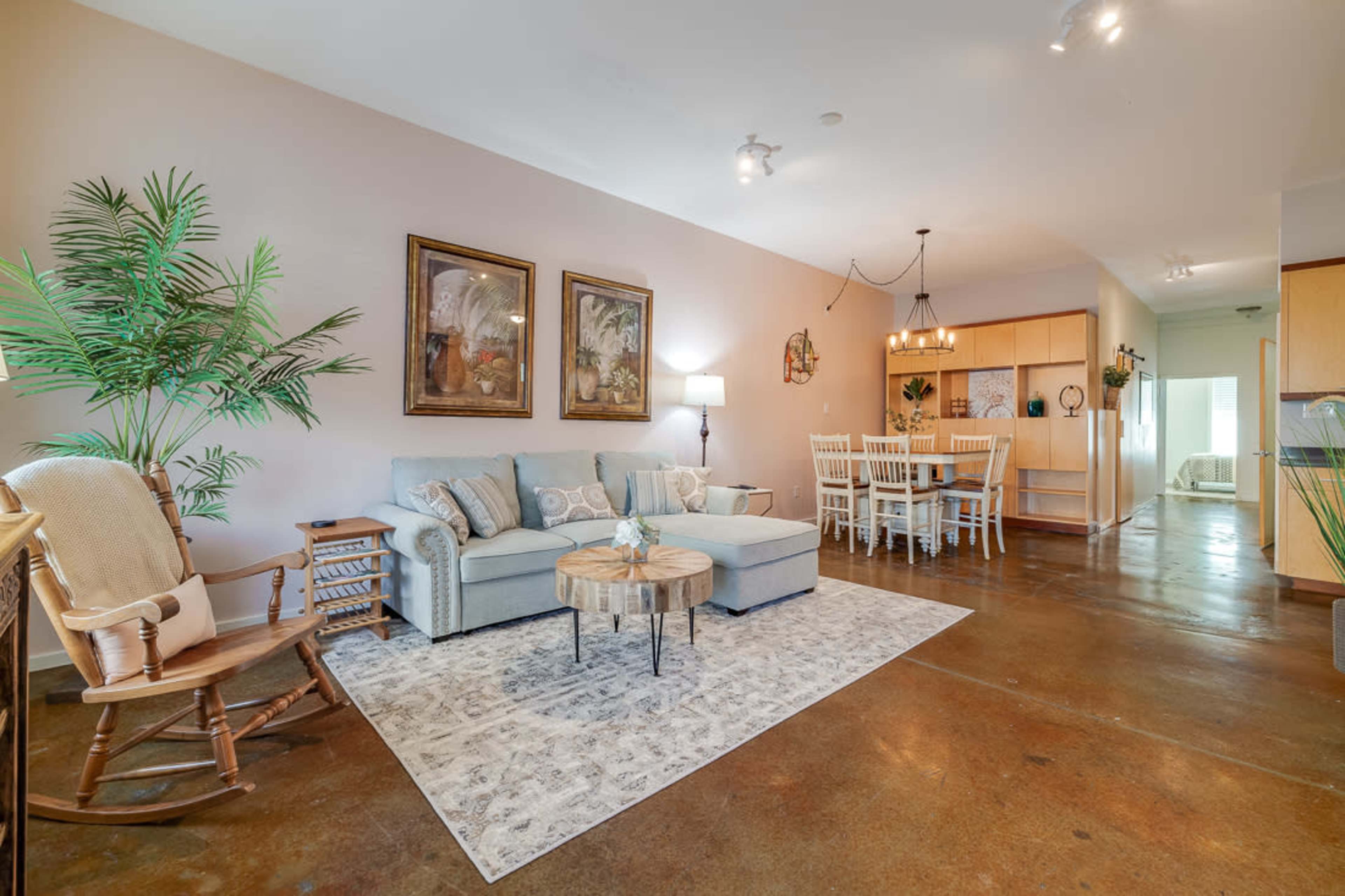 The image shows a living room with a light blue sectional sofa, a round coffee table, and a dining area in the background featuring a rectangular table and chairs against a neutral-colored wall.