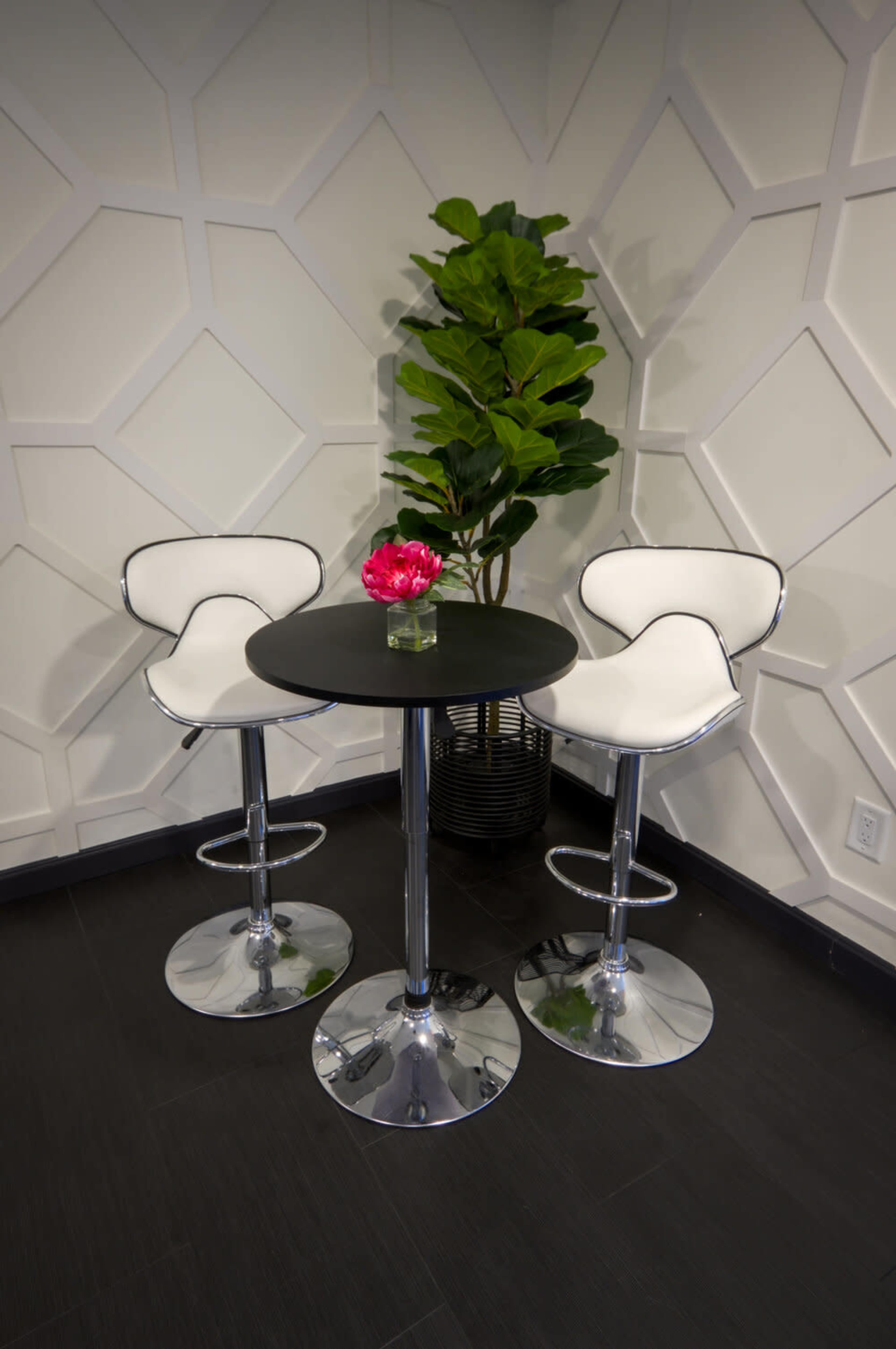 A modern black table with two white bar stools and a small pink flower arrangement stands beside a leafy green plant against a textured white wall.
