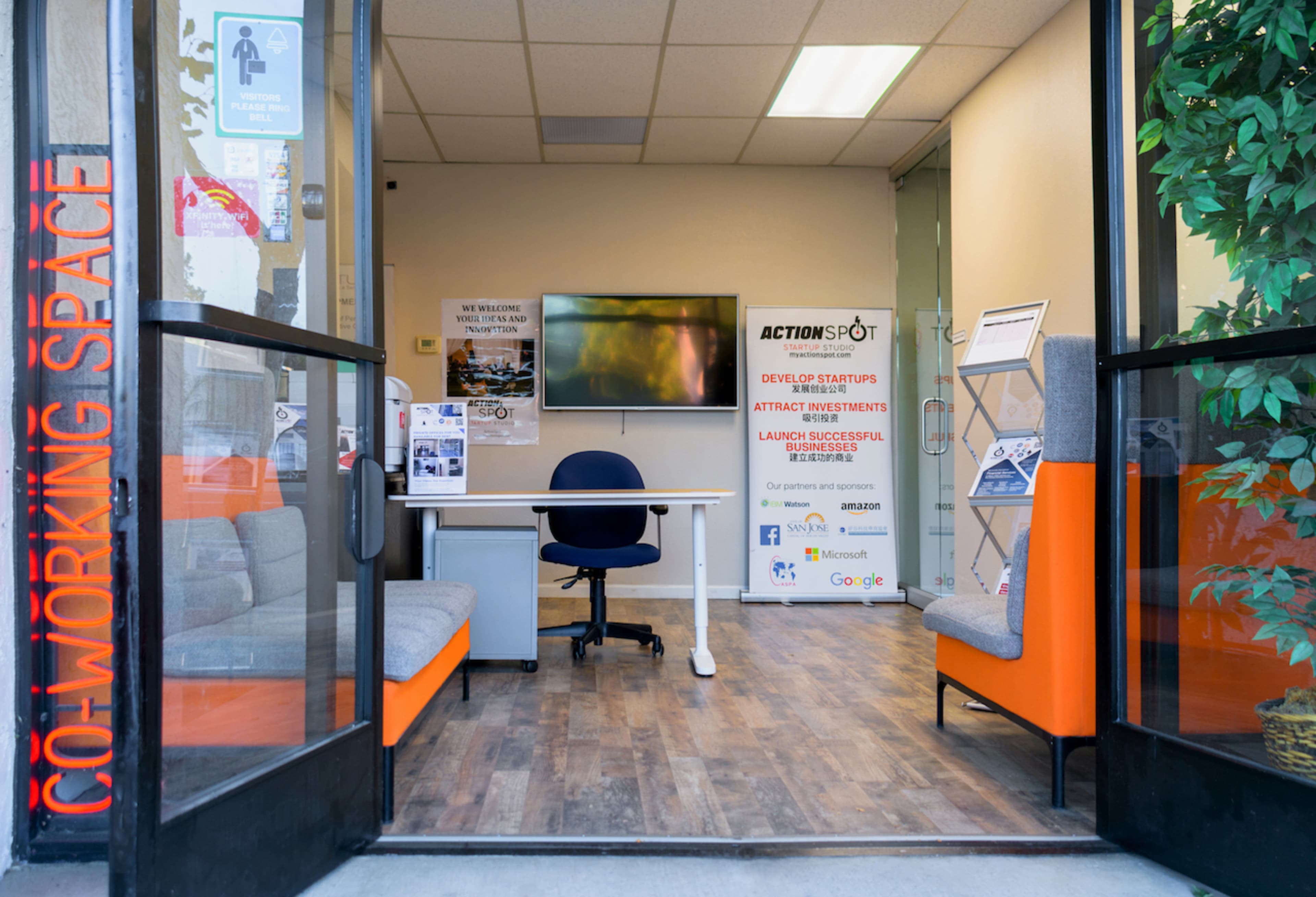 The entrance of a co-working space featuring a modern desk and seating area, with a monitor displayed on the wall and various informational posters around.