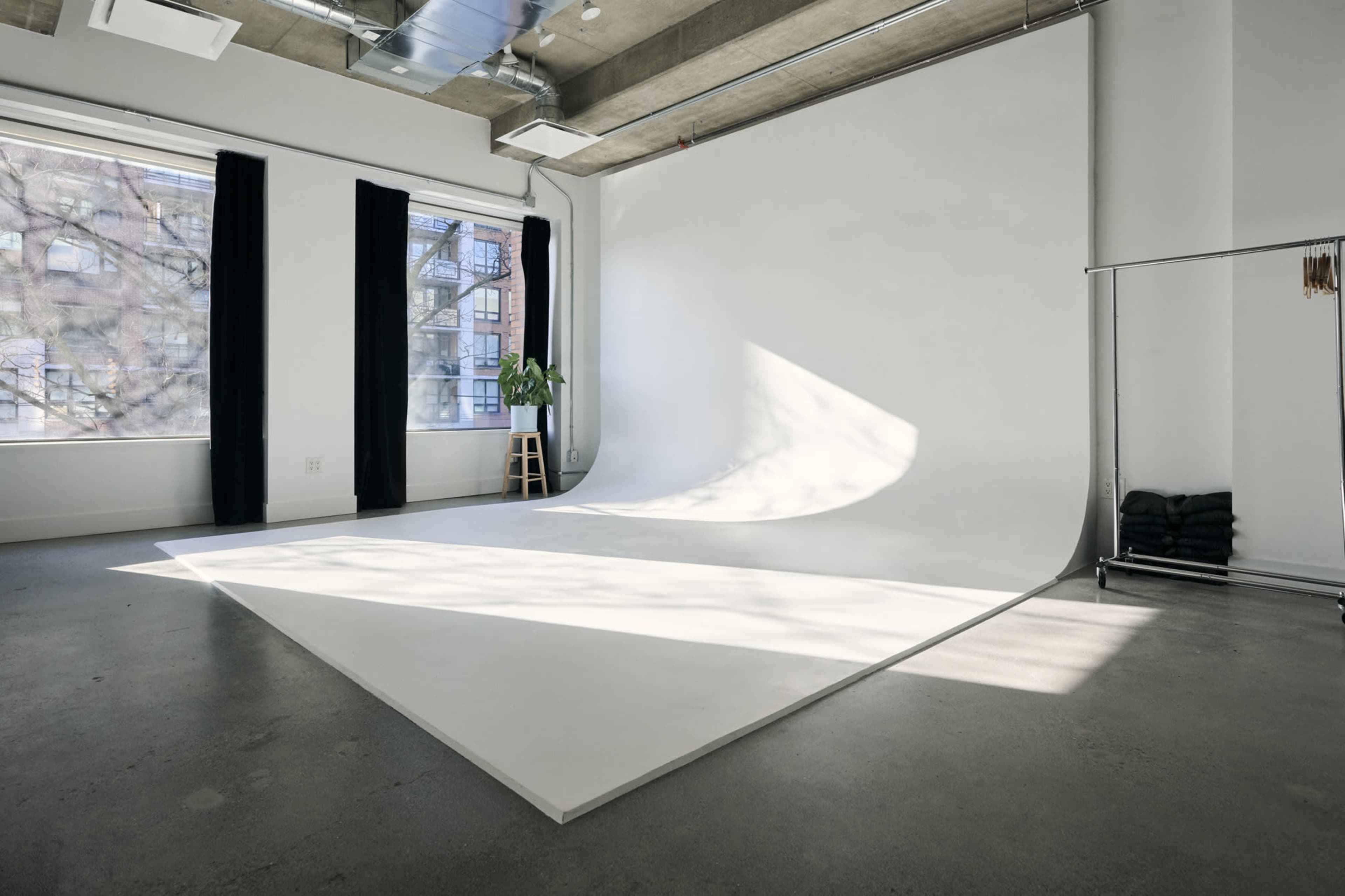 The image shows a spacious photography studio with a large white backdrop, floor-to-ceiling windows, and a plant in the corner.