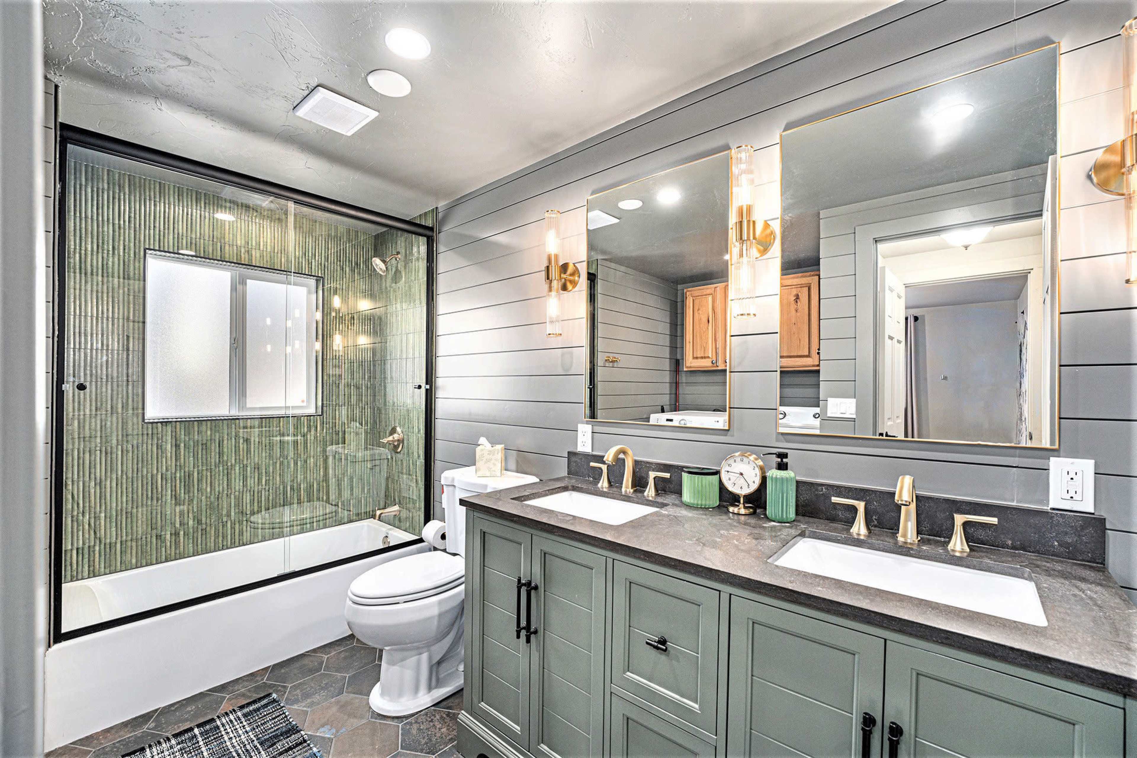The image shows a modern bathroom featuring a glass shower enclosure, a bathtub, dual sinks with brass fixtures, and a combination of green cabinetry and hexagonal tile flooring.