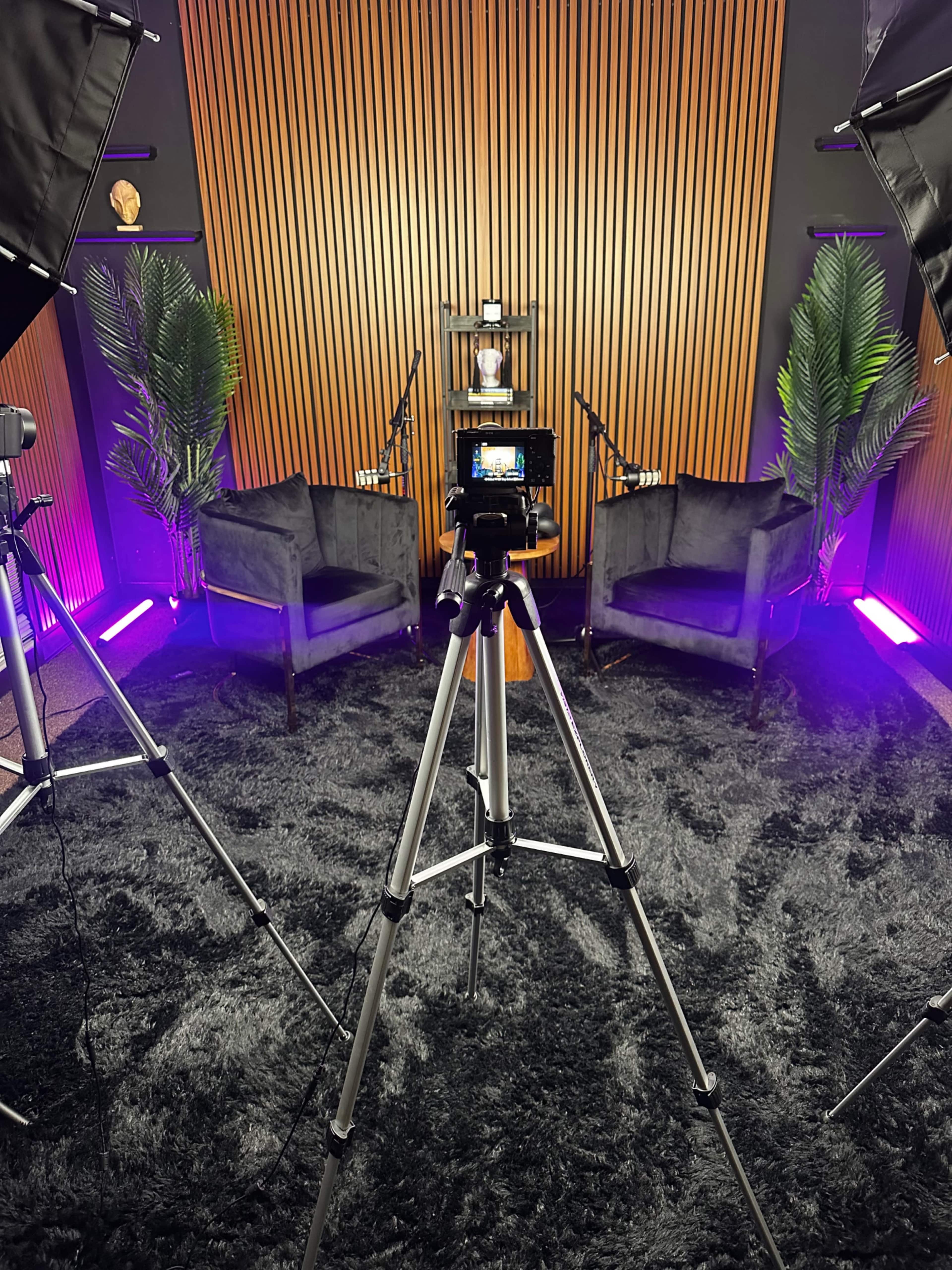 The image shows a well-lit recording studio setup with two chairs, a camera on a tripod, and ambient lighting.