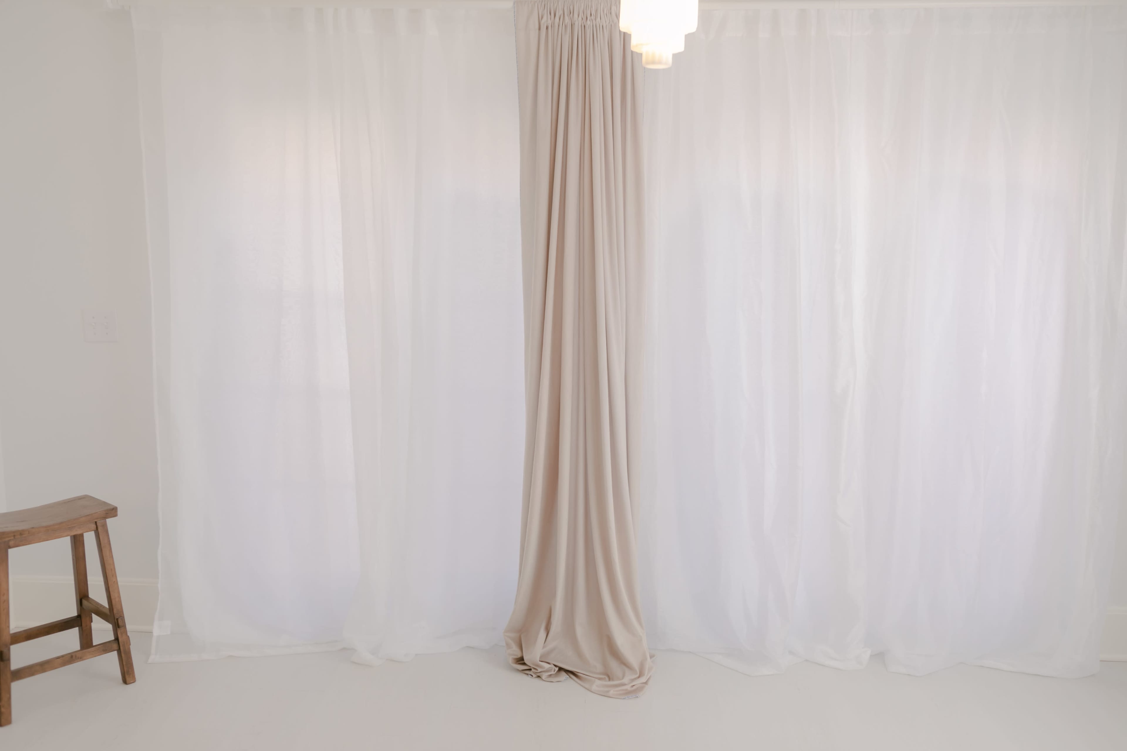 A room with sheer white curtains and a beige curtain hanging from the ceiling, alongside a wooden stool.