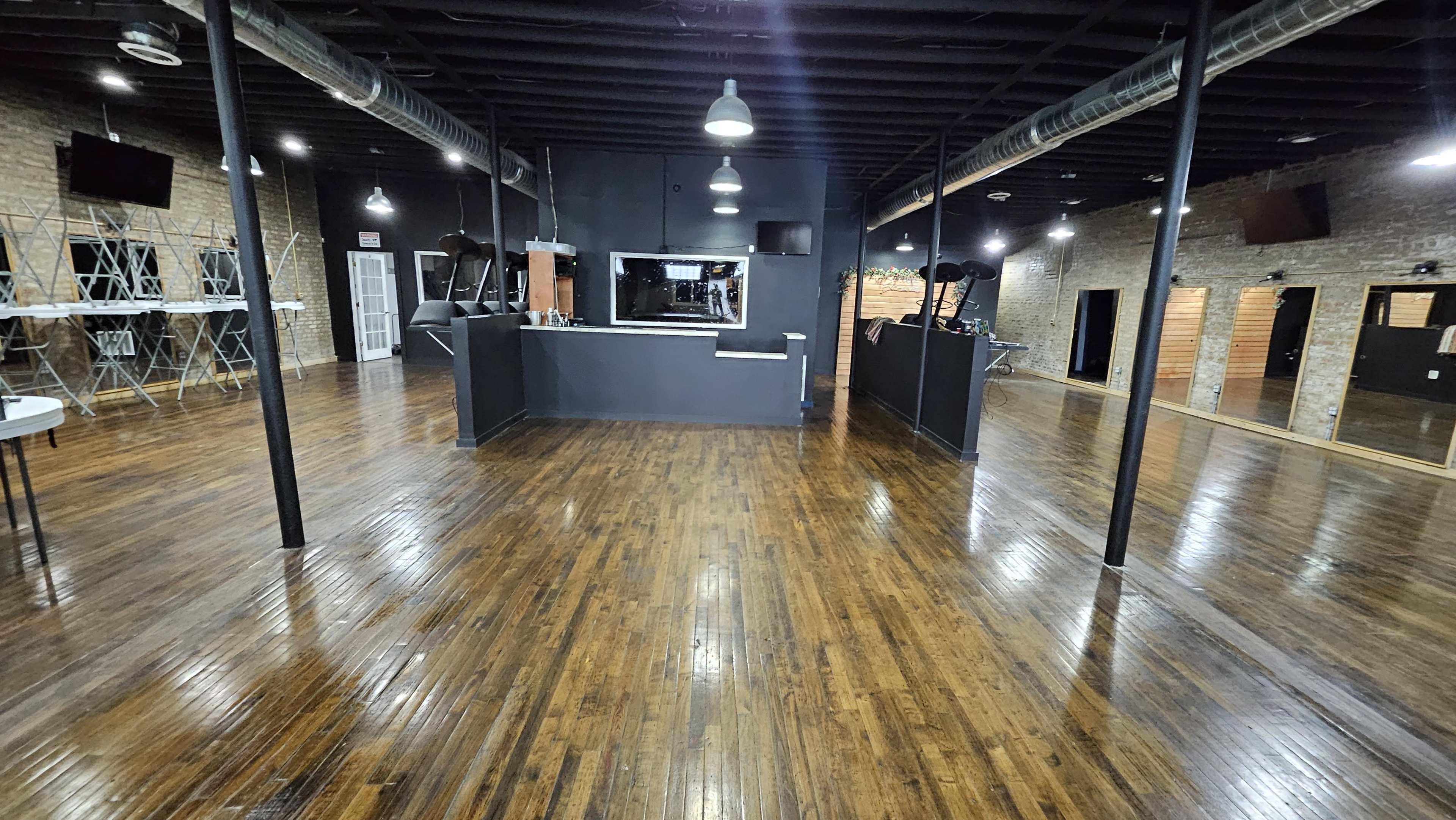 The image shows a spacious commercial interior with polished wooden floors, exposed brick walls, and multiple television screens mounted on the walls.