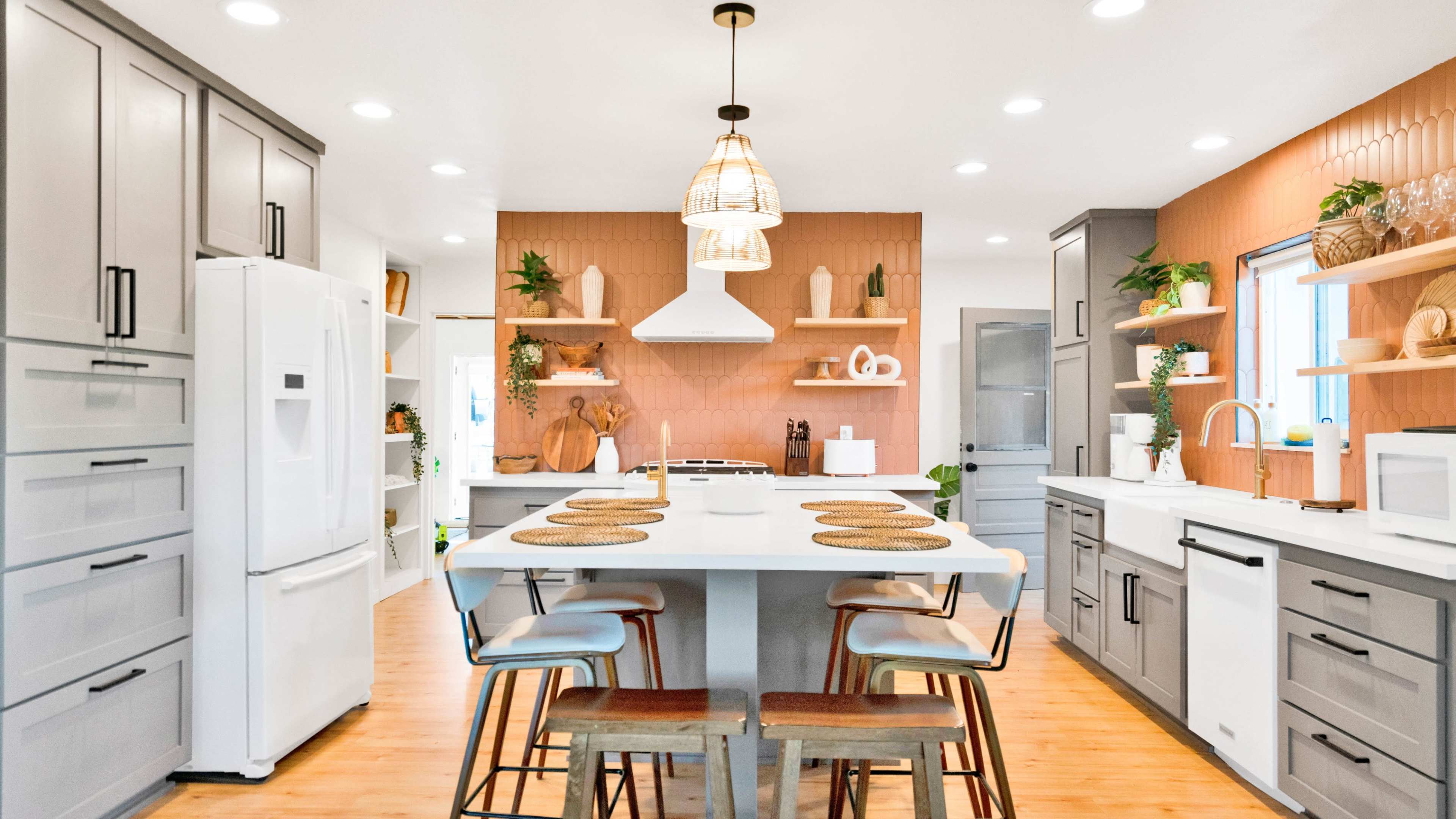 A modern kitchen features gray cabinets, a central island with wooden stools, and warm-toned accent walls adorned with plants and decorative shelves.