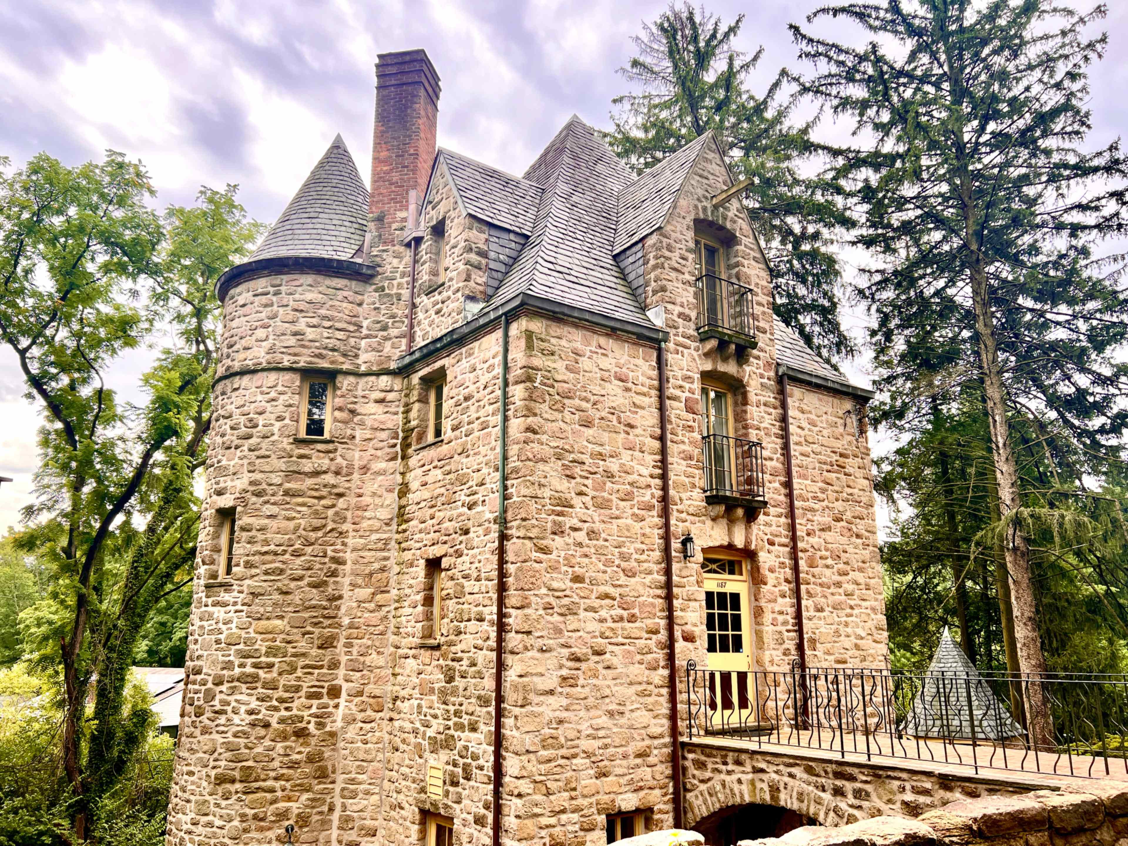 A stone castle with a steeply pitched roof and multiple towers is surrounded by tall trees.