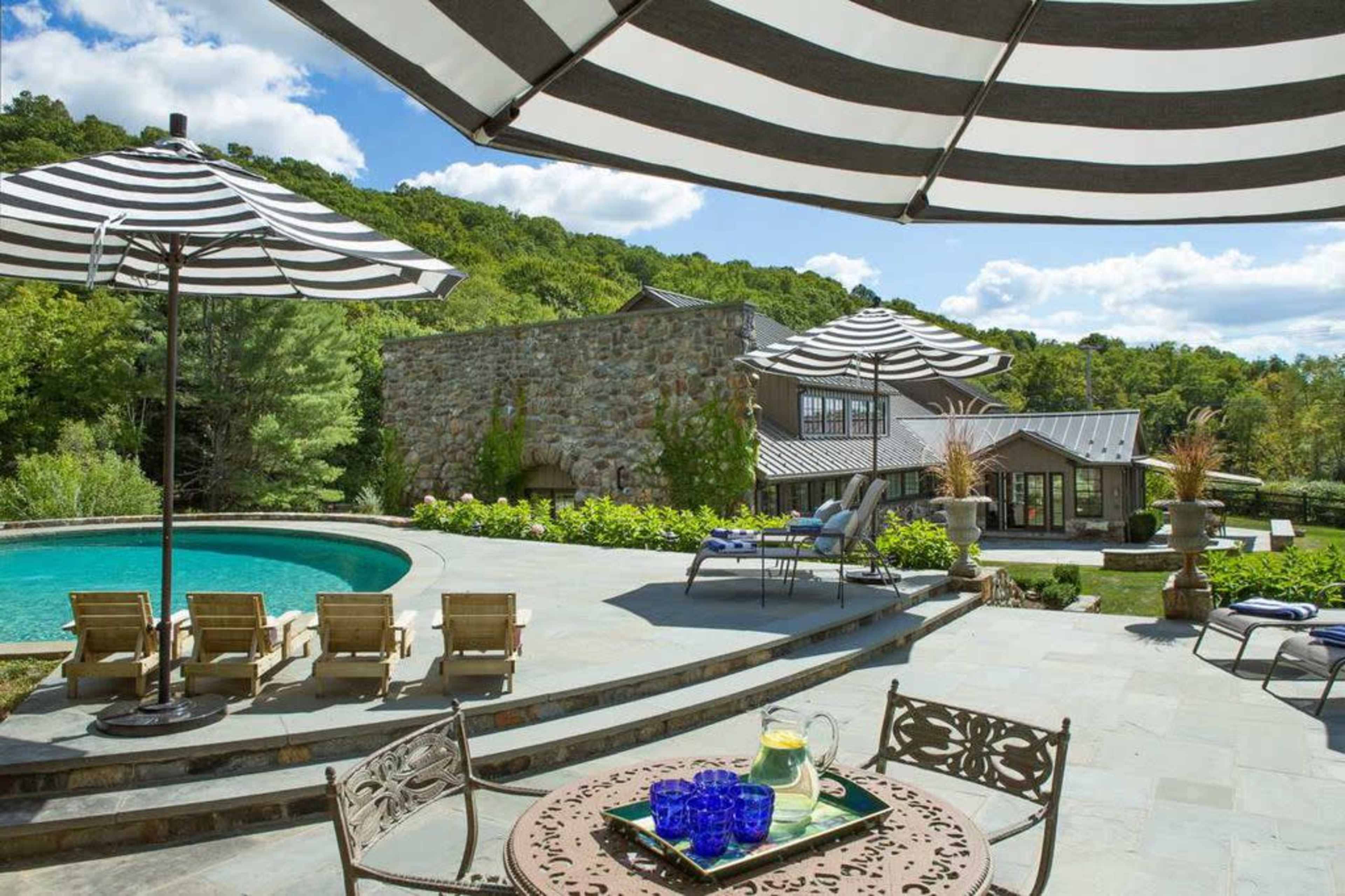 The image shows a well-designed outdoor space featuring a pool, lounge chairs, and striped umbrellas, with a stone wall and a house in the background surrounded by greenery.