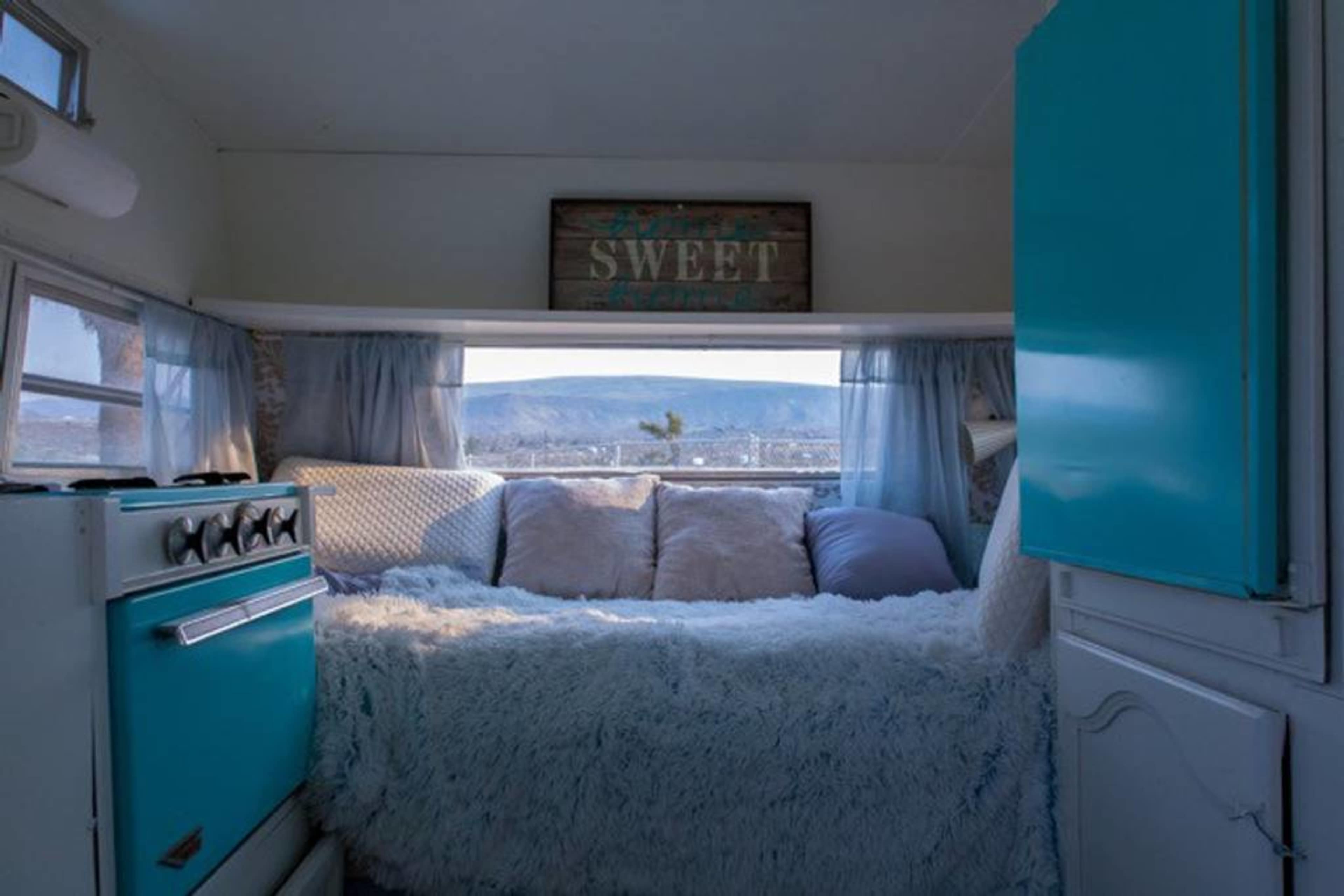 The interior of a small camper features a turquoise kitchen area, a cozy bed with soft cushions, and a window showcasing a mountain view.