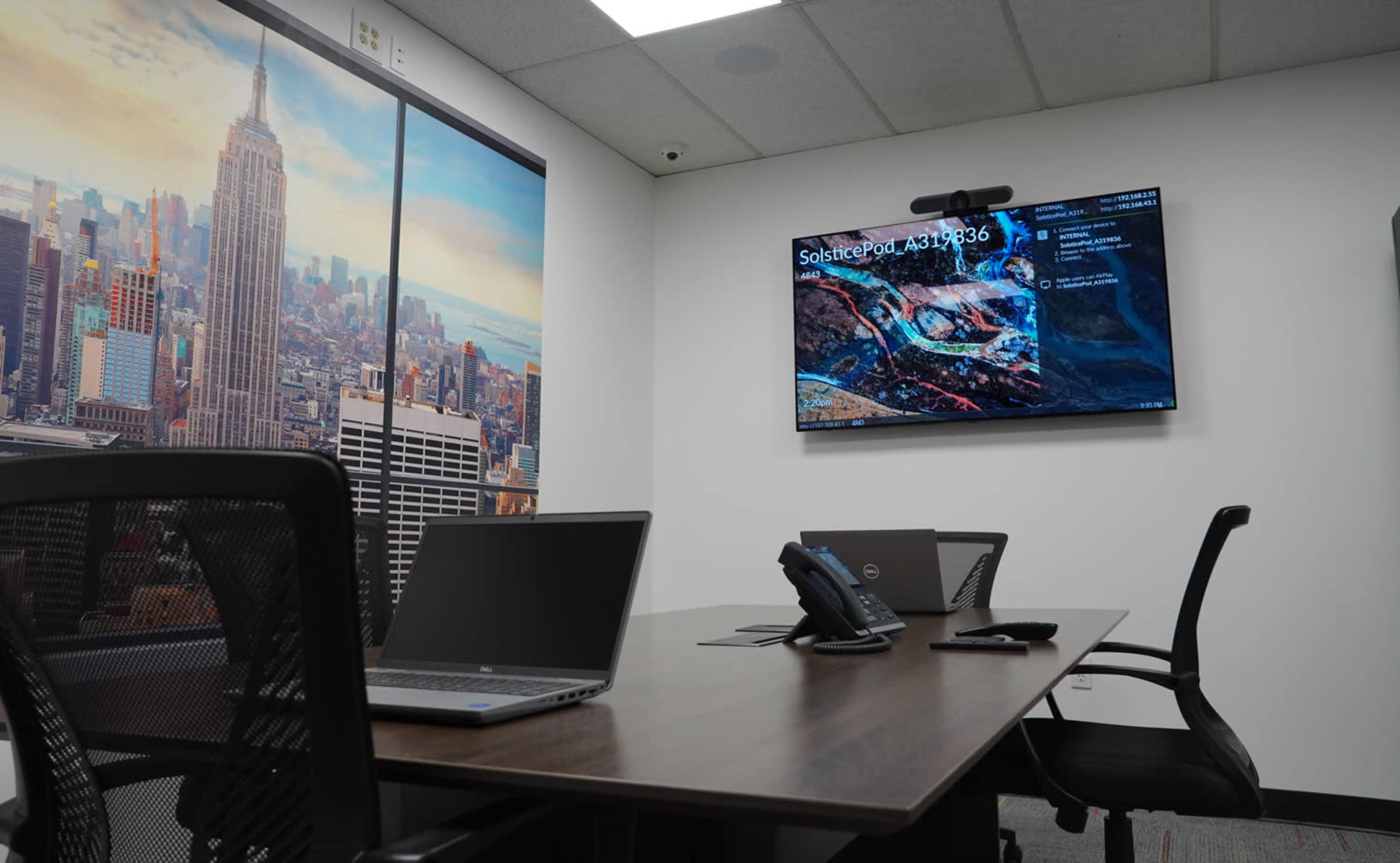 A conference room features a large screen displaying a presentation, laptops on a wooden table, and a wall decorated with a city skyline.