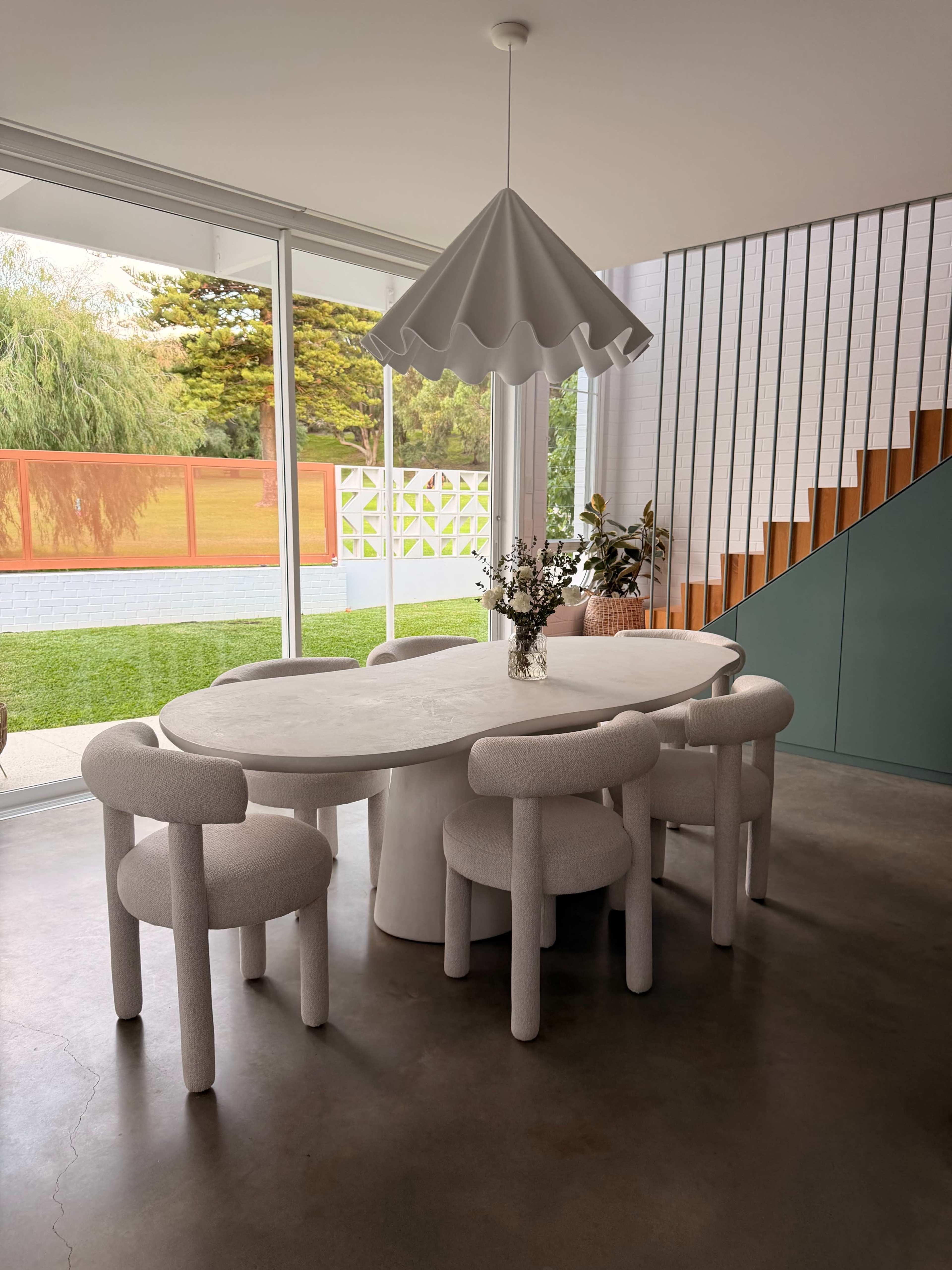 A modern dining area features a round table surrounded by four rounded chairs, with a pendant light hanging above and a view of a green outdoor space through large glass windows.