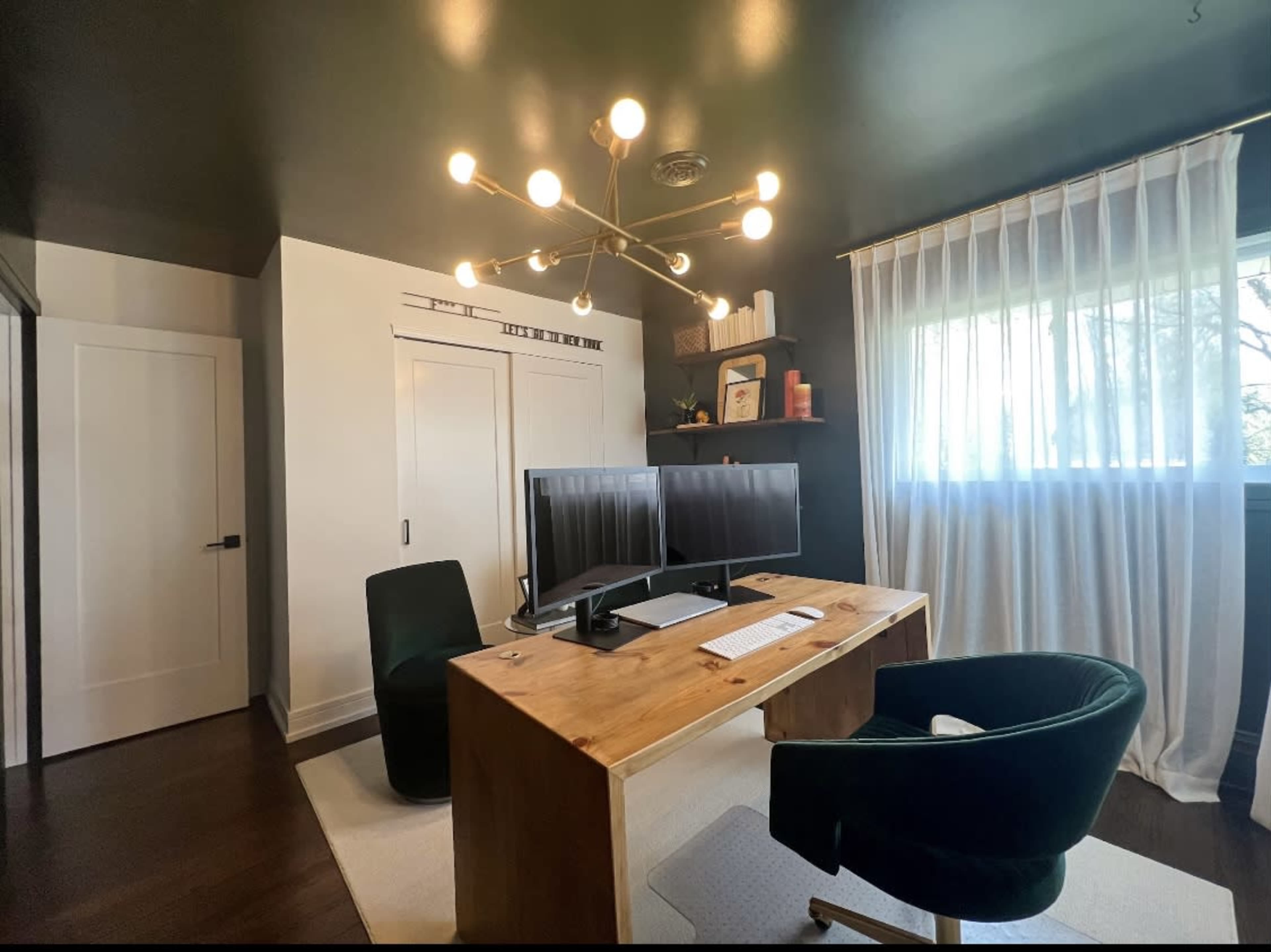 A modern home office with a wooden desk, dual monitors, a green chair, and large windows covered by sheer white curtains.