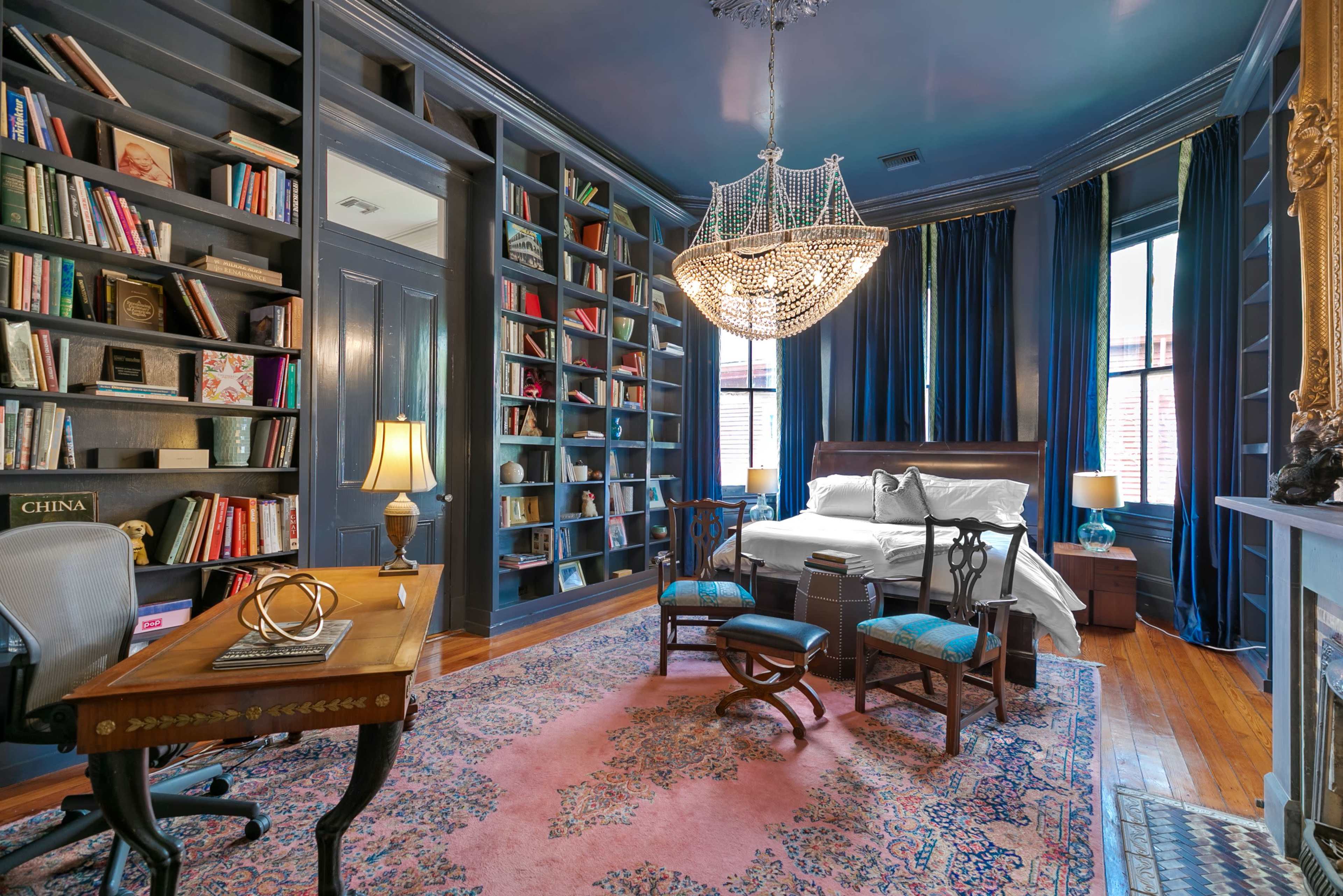 The image depicts a bedroom with dark blue walls, a chandelier, a bookshelf filled with books, a desk, and a patterned rug.