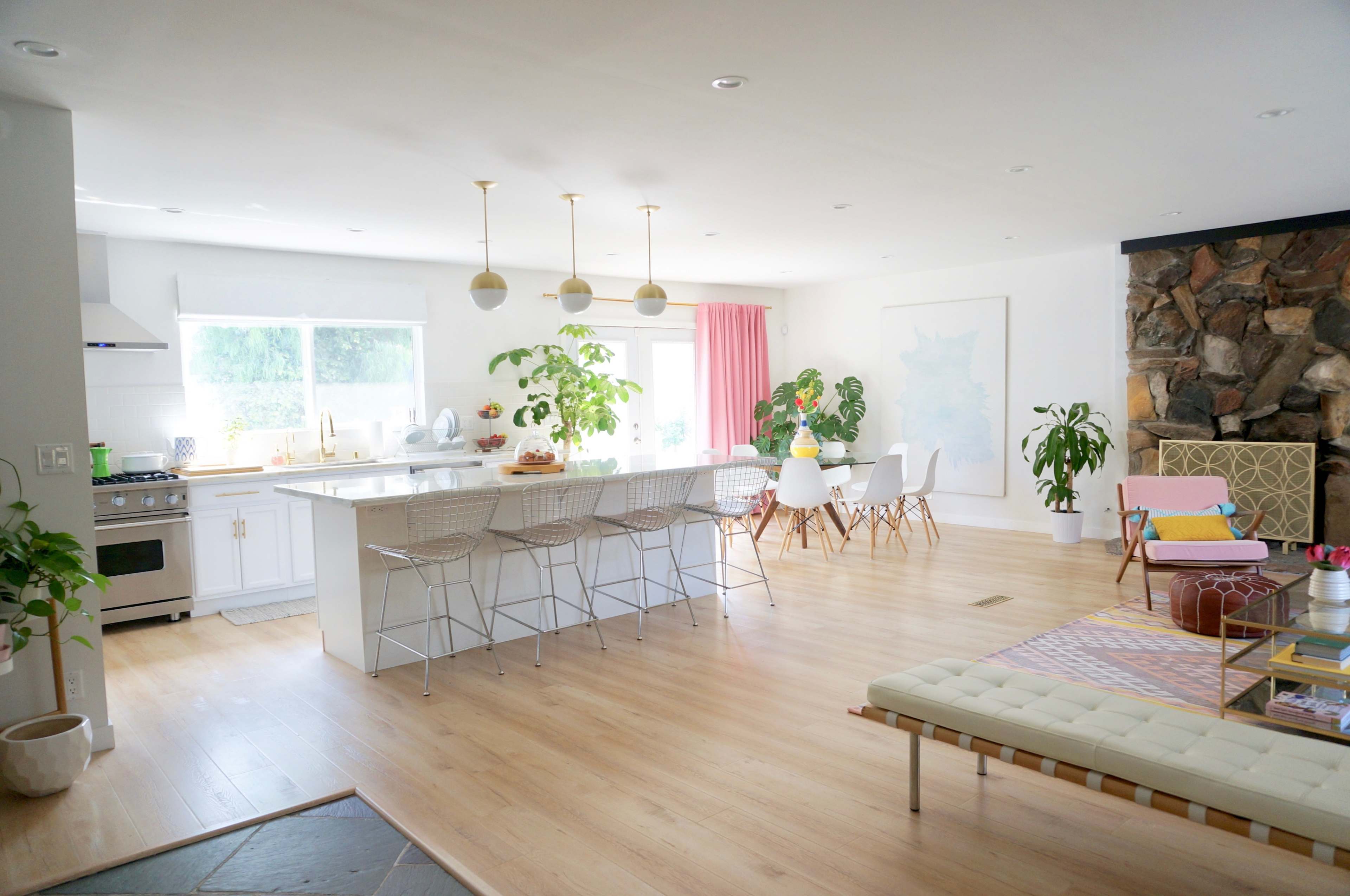 A bright, modern kitchen and dining area features a large island with bar stools, white cabinetry, and a stone fireplace, complemented by plants and colorful furnishings.