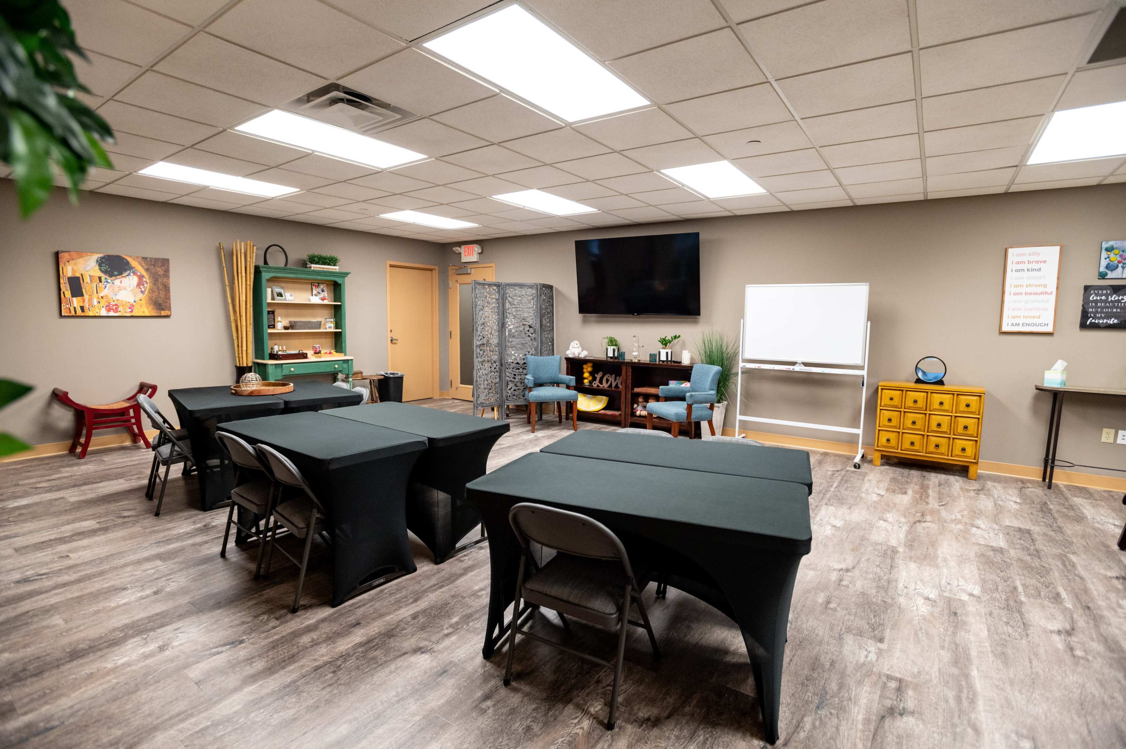 A modern training room is arranged with several black tables and chairs, featuring a TV, a whiteboard, and decoration along the walls.