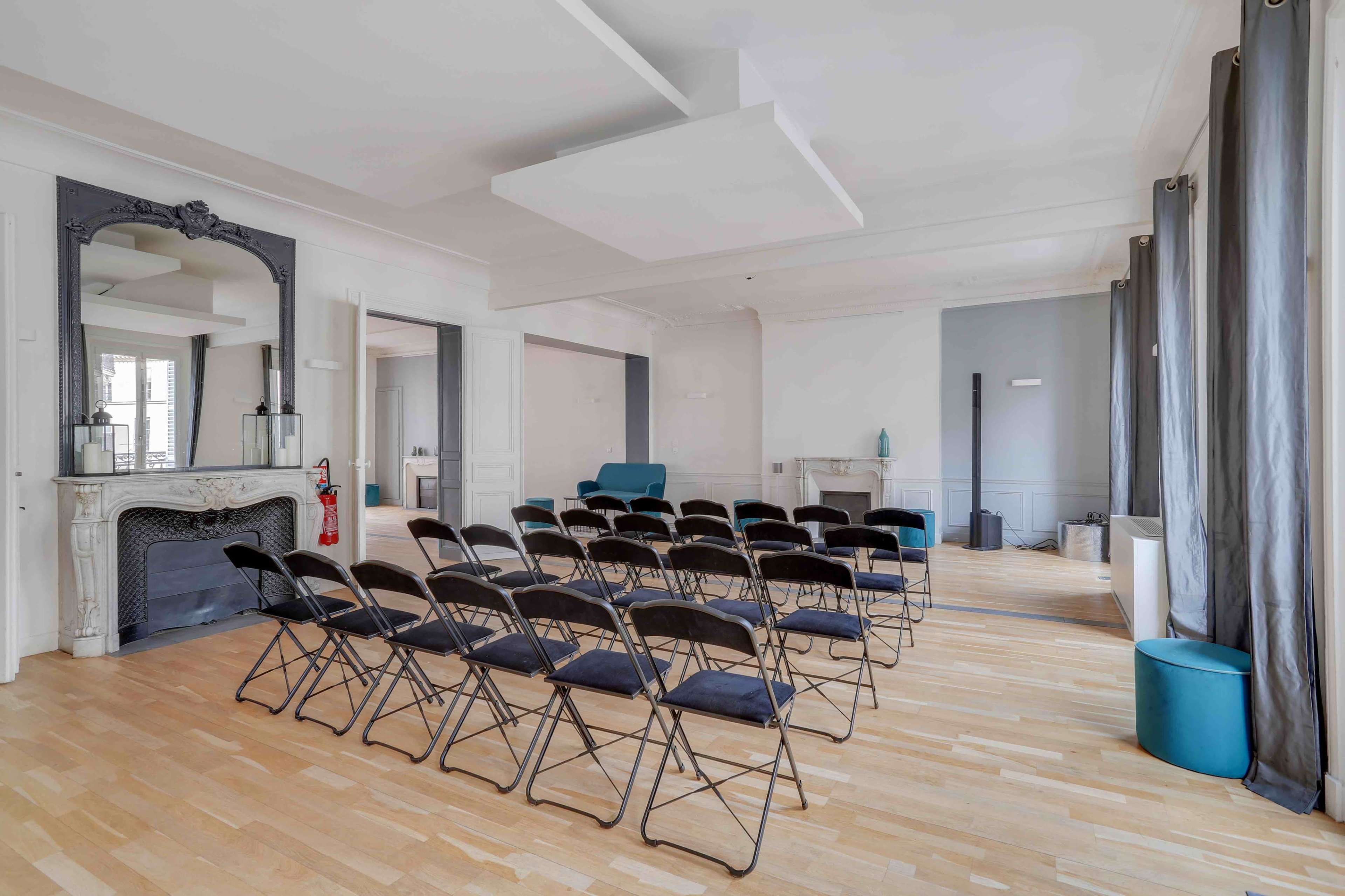 A spacious room with a row of black folding chairs arranged facing a light blue wall, with a fireplace and large windows dressed in gray curtains.