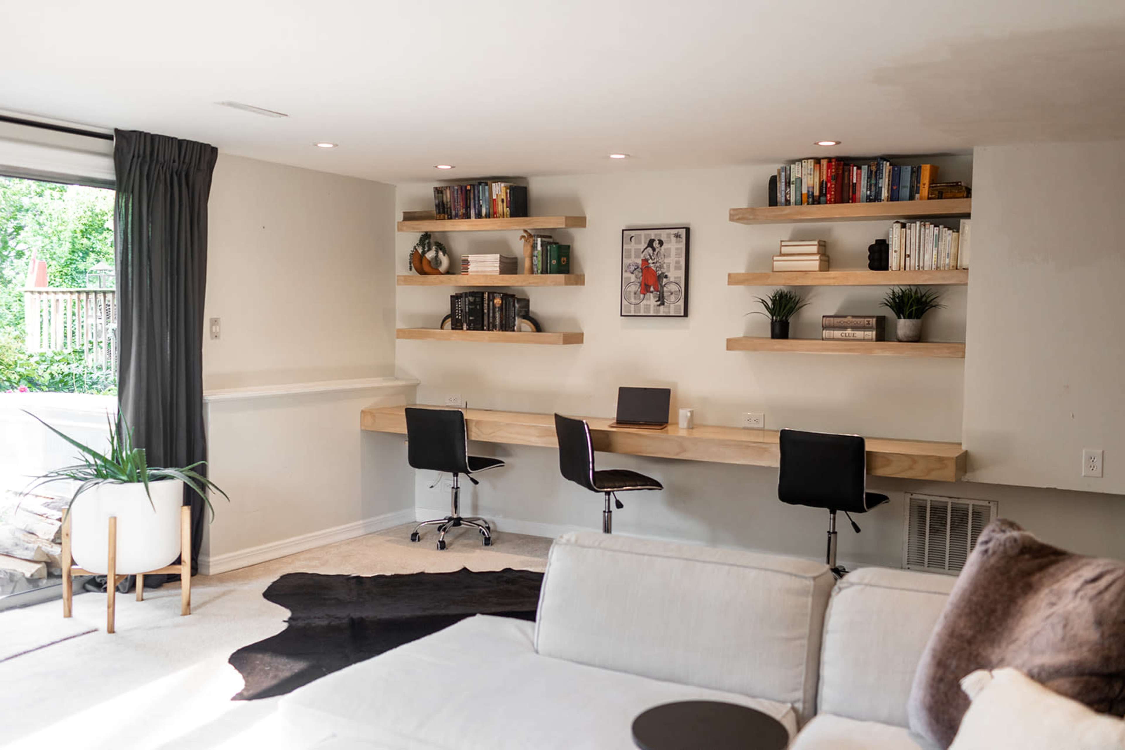 A minimalist home office features a long wooden desk with two chairs, surrounded by wall-mounted shelves filled with books and decorative plants.