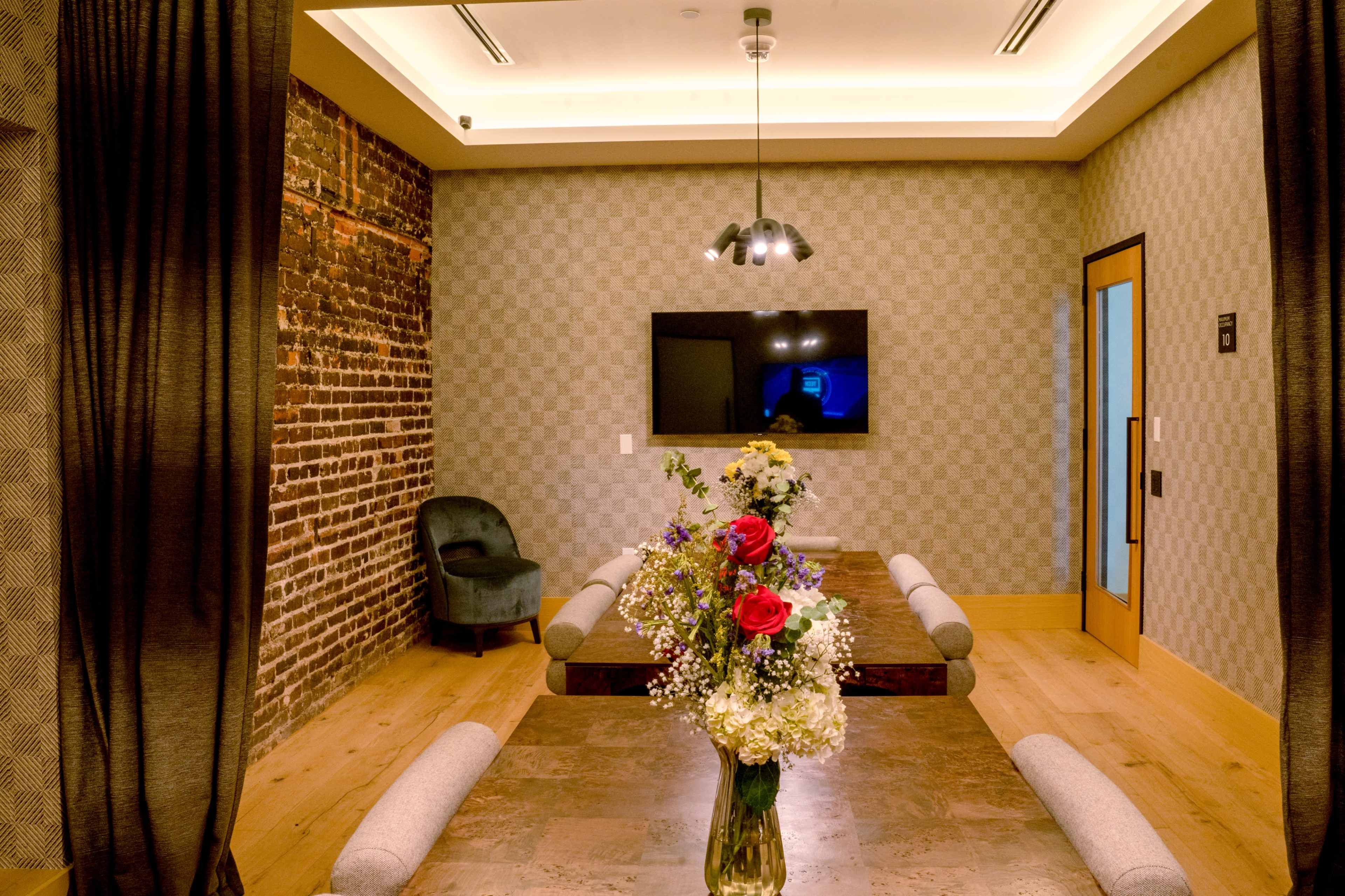 A modern meeting room features a wooden table surrounded by upholstered benches, a floral centerpiece, a television on the wall, and exposed brick accents.