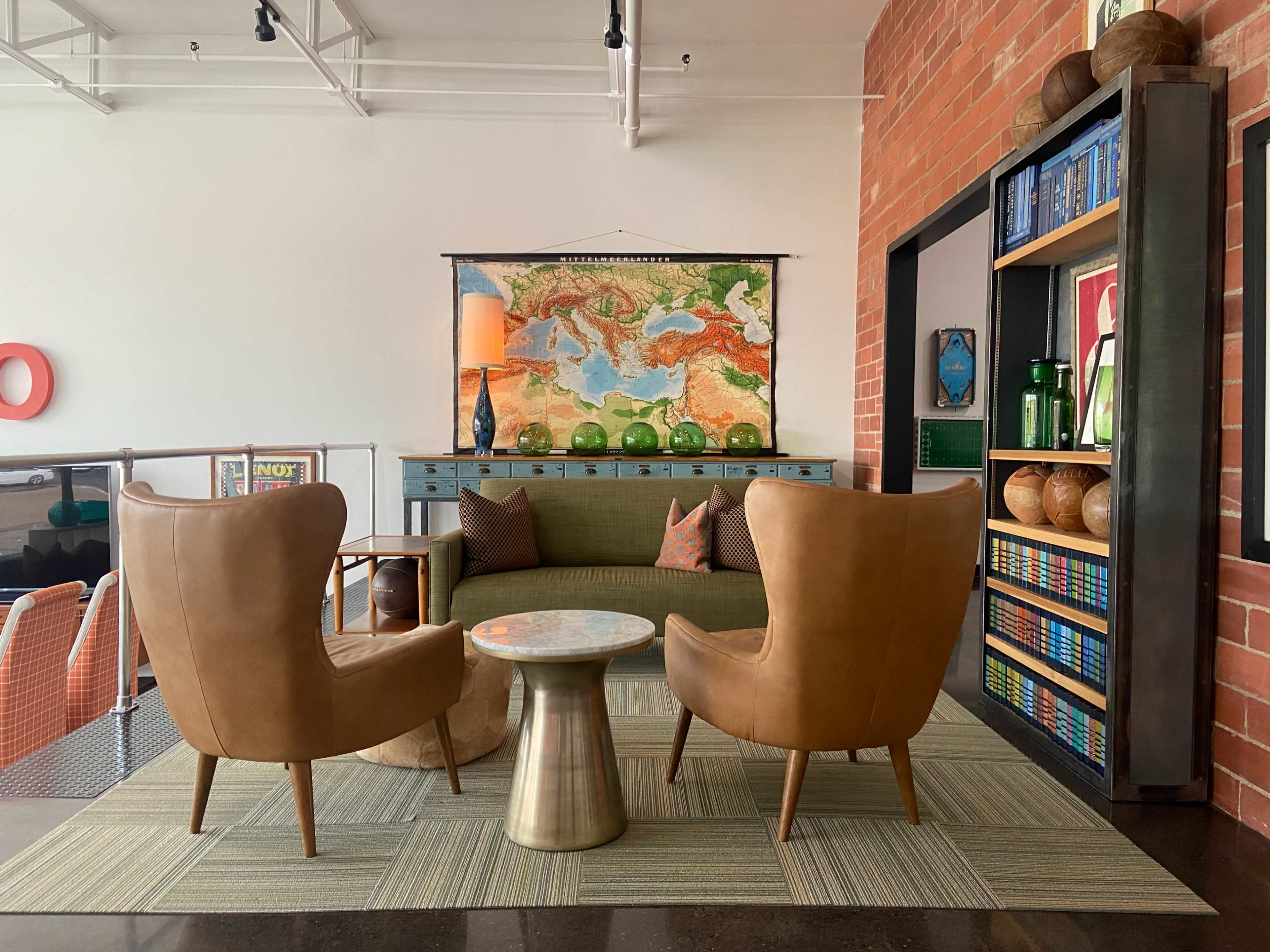 A modern seating area with two brown armchairs facing a small round table, framed by a colorful map on the wall and shelves filled with decorative items.
