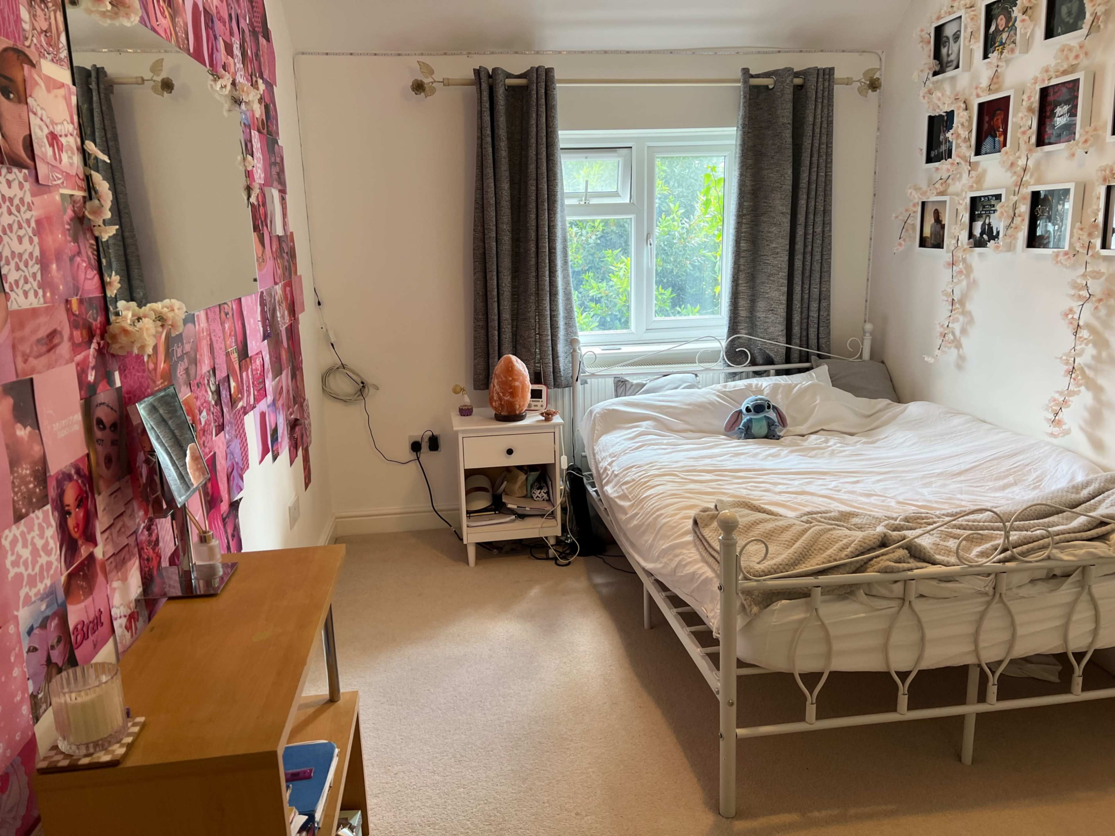 The room features a bed with white bedding, a nightstand, a wooden dresser, and walls decorated with pink photographs and curtains covering the window.