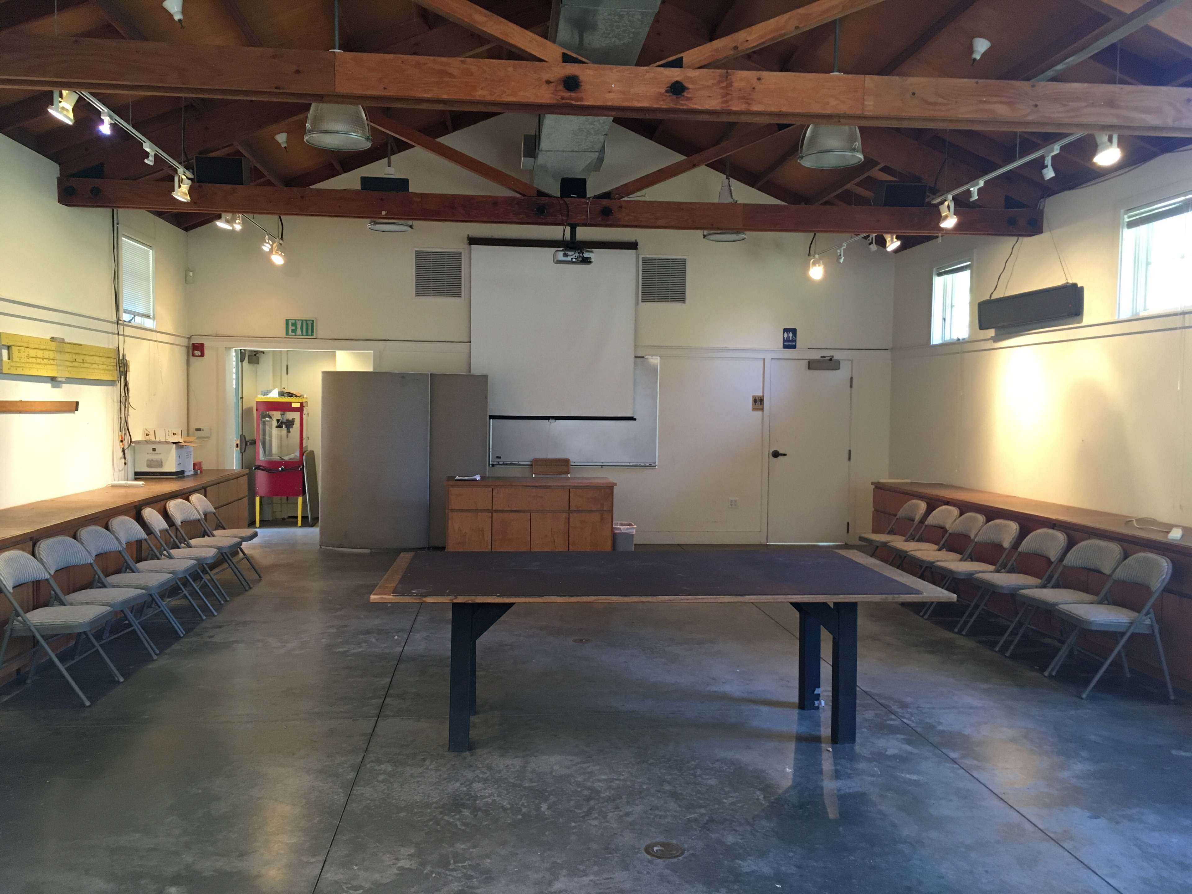 A spacious room with a wooden ceiling features a central table, chairs arranged along the walls, and a projection screen at the front.
