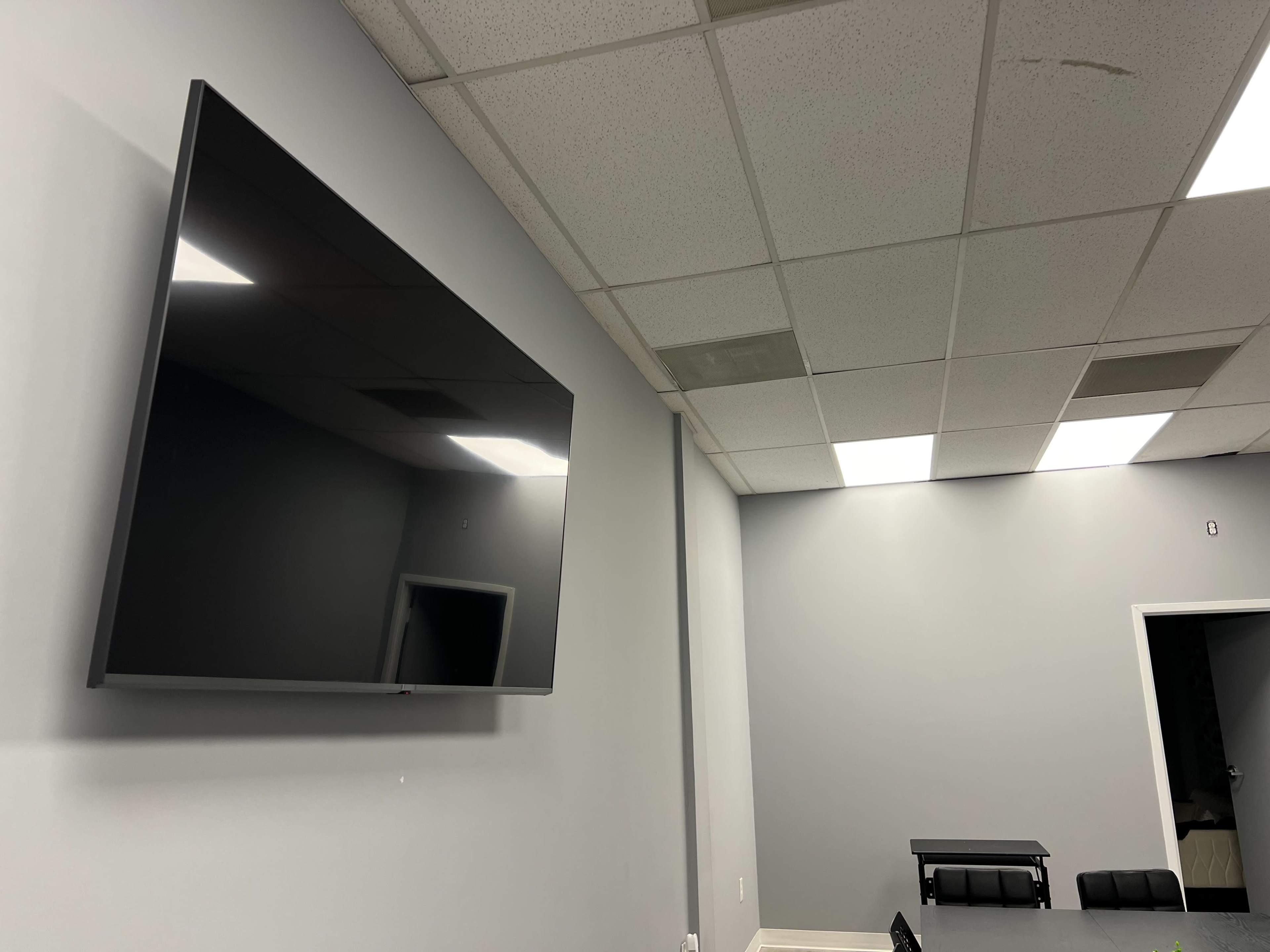 A wall-mounted flat-screen television is installed in a gray meeting room with fluorescent ceiling lights.