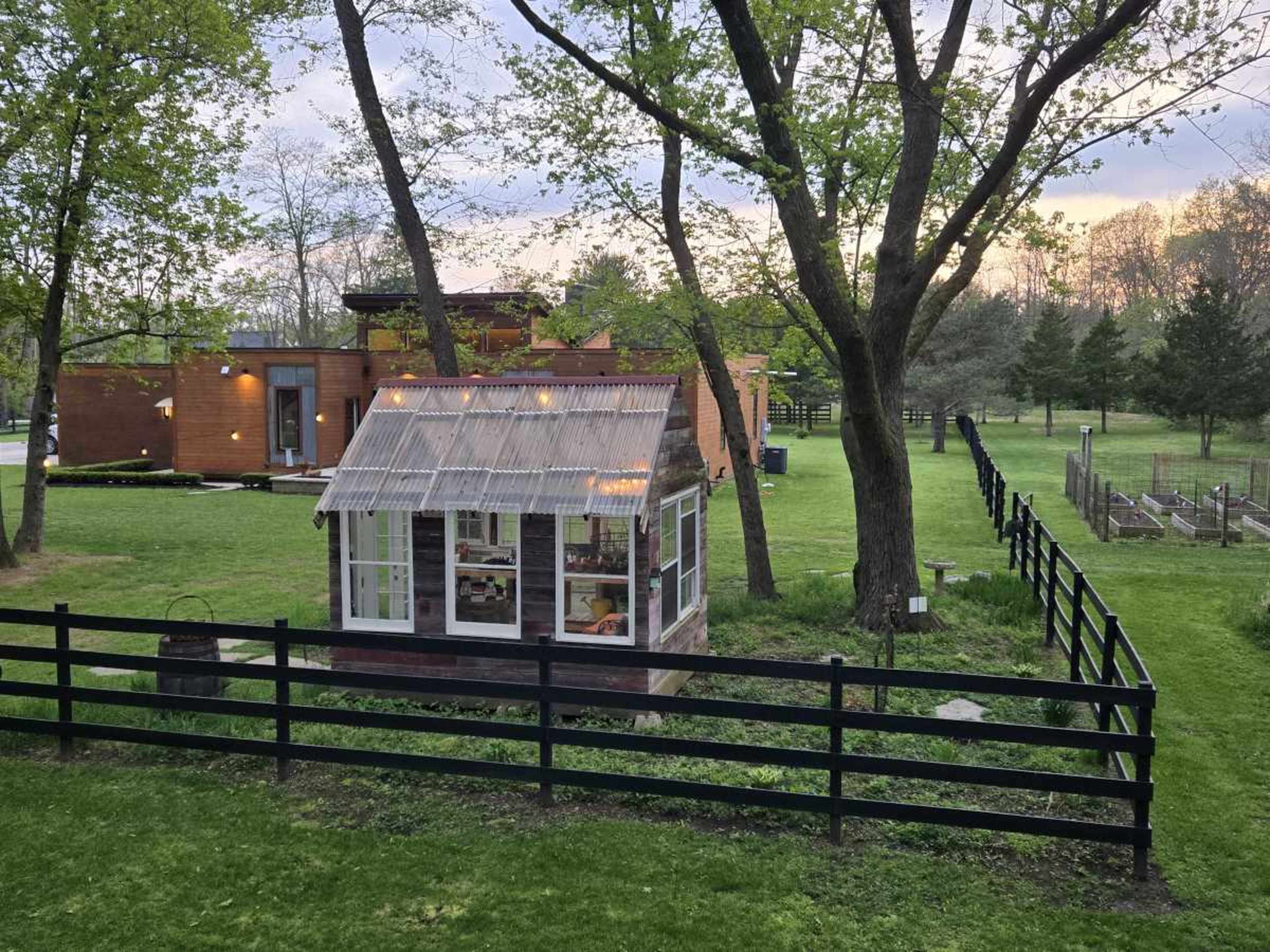 The image shows a small greenhouse made of glass and wood amidst a grassy area, with a modern house and a fenced garden in the background.