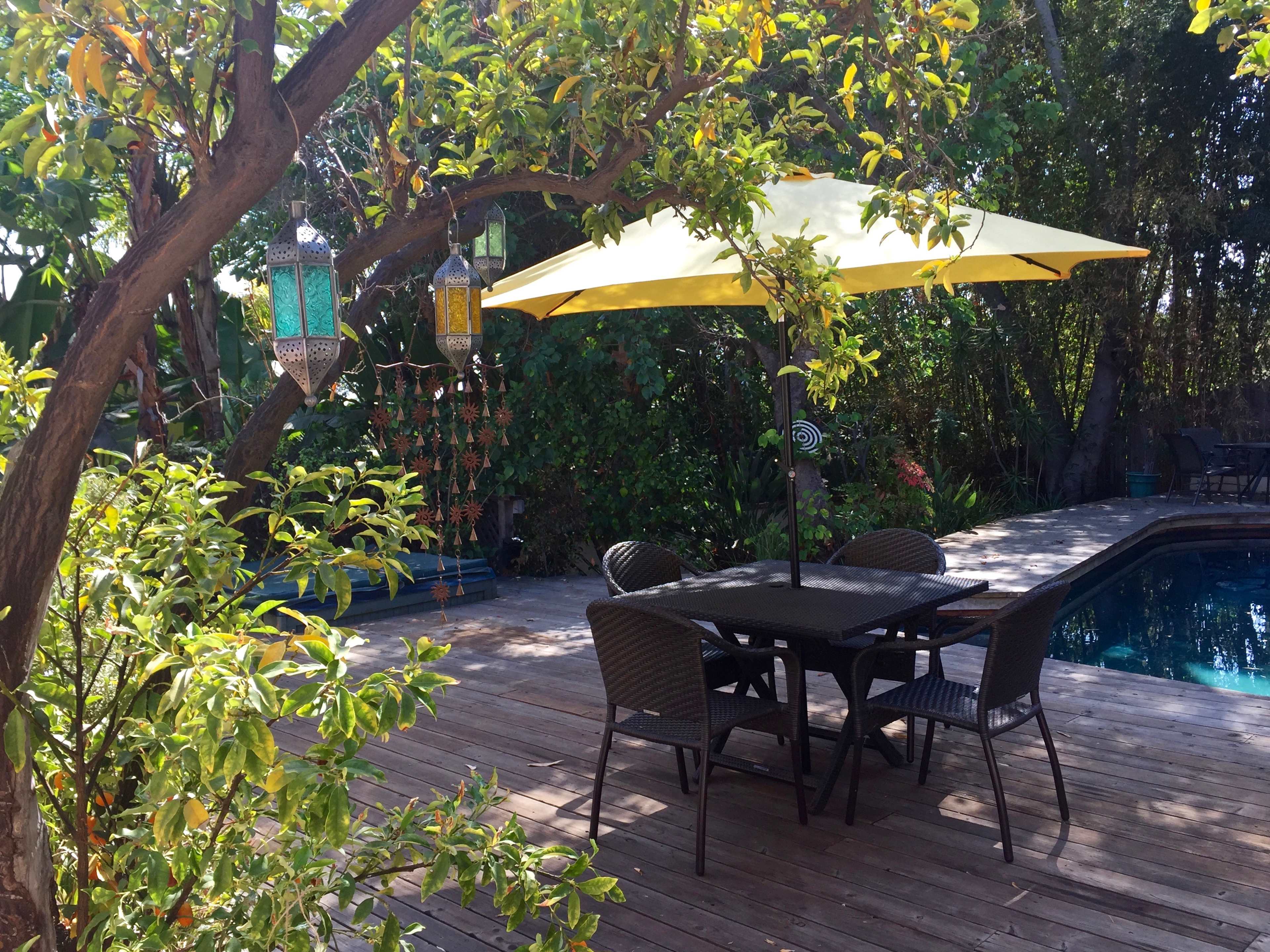 A patio with a black dining table and chairs sits beneath a yellow umbrella, surrounded by greenery and a swimming pool.