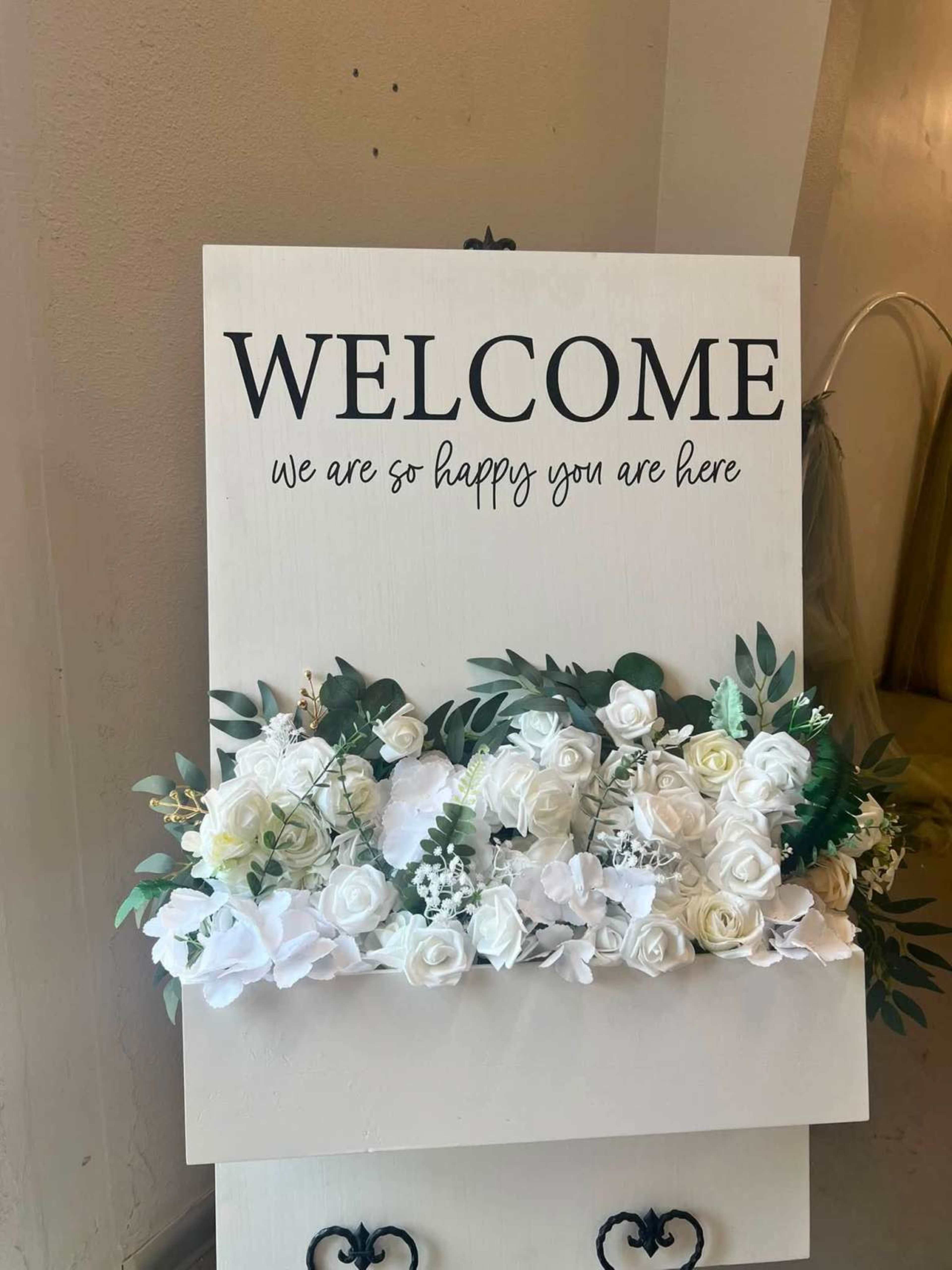 A welcome sign adorned with white and green artificial flowers is displayed on a stand.