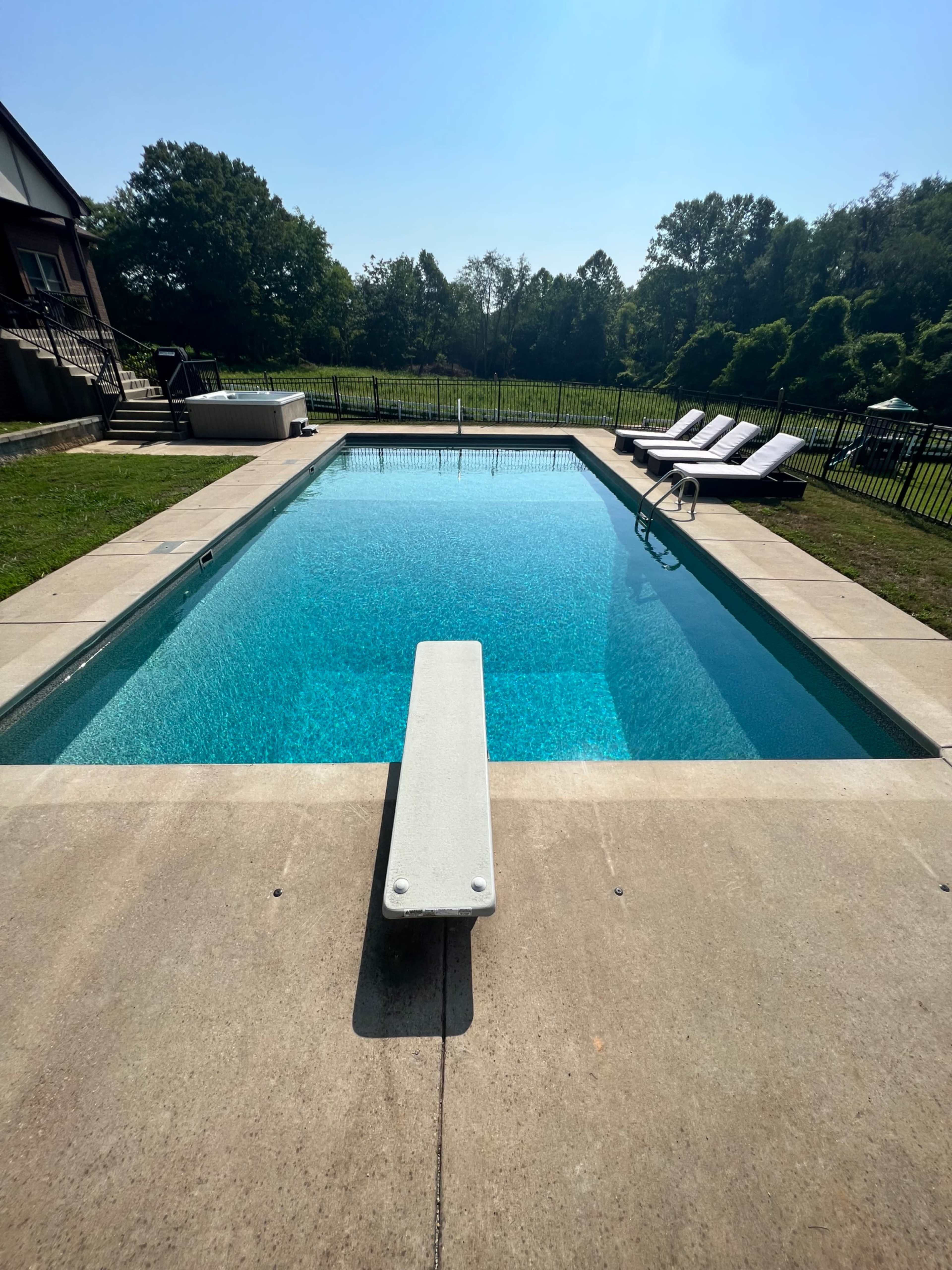 The image shows a rectangular swimming pool with a diving board at one end, surrounded by a concrete deck and lounge chairs under a clear blue sky.