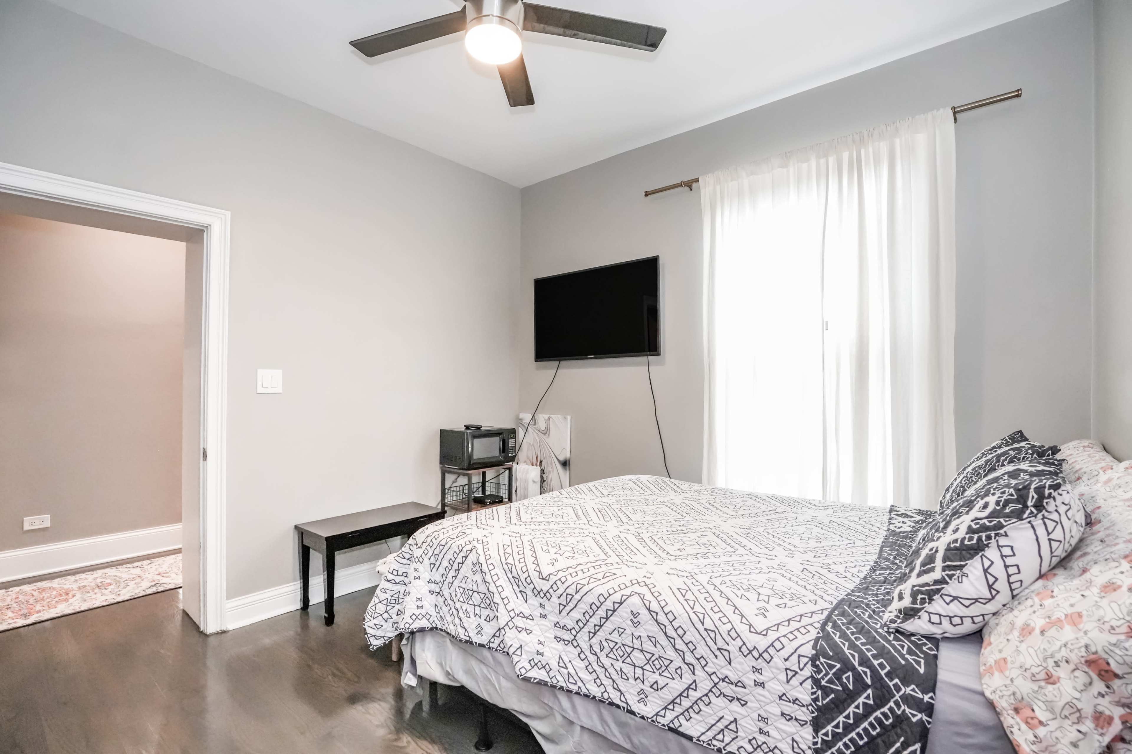 A bedroom with a bed covered in a patterned blanket, a television mounted on the wall, and a window with sheer curtains.