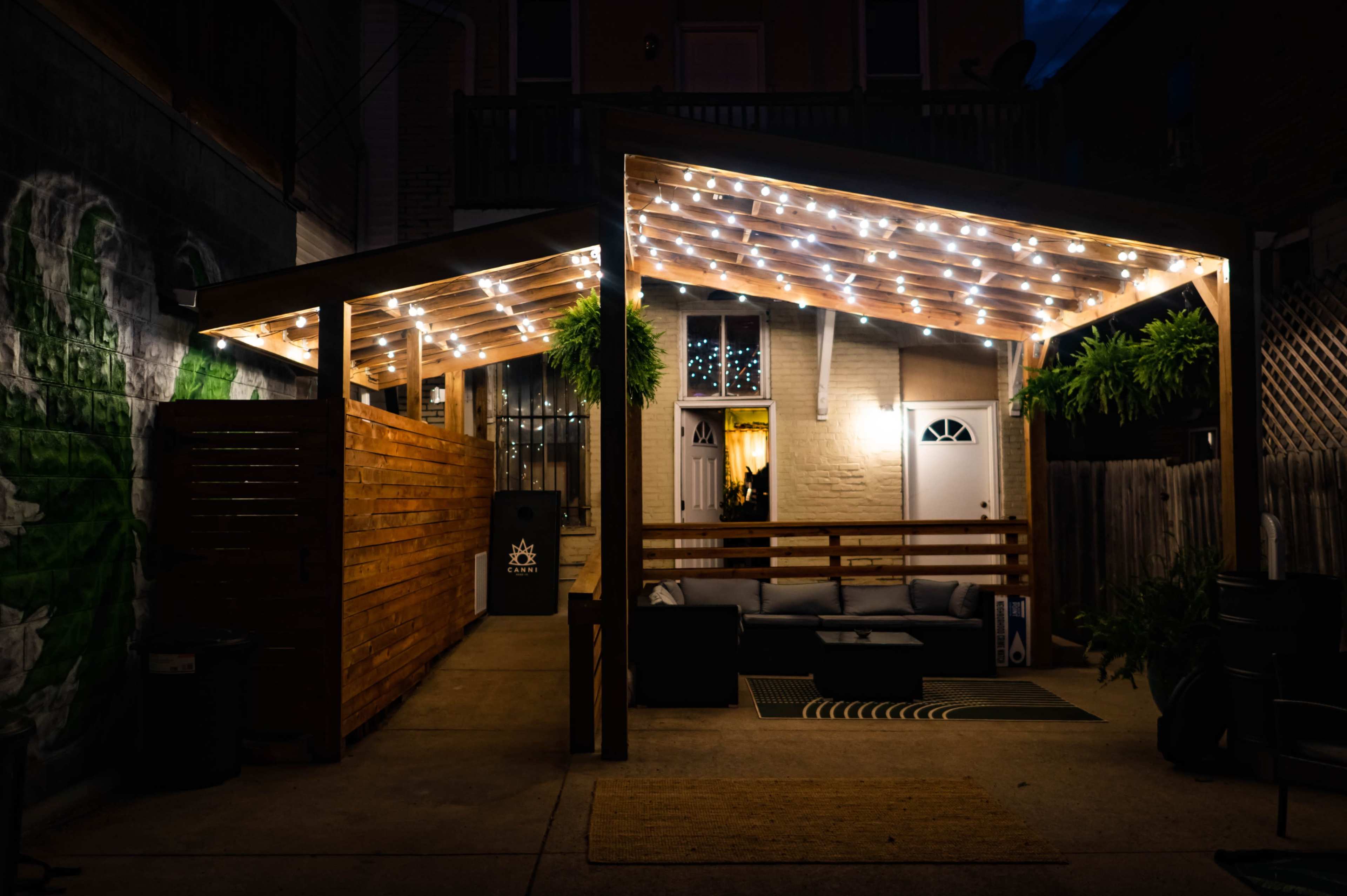A cozy outdoor patio with string lights and a sectional sofa is positioned beneath a wooden roof structure.