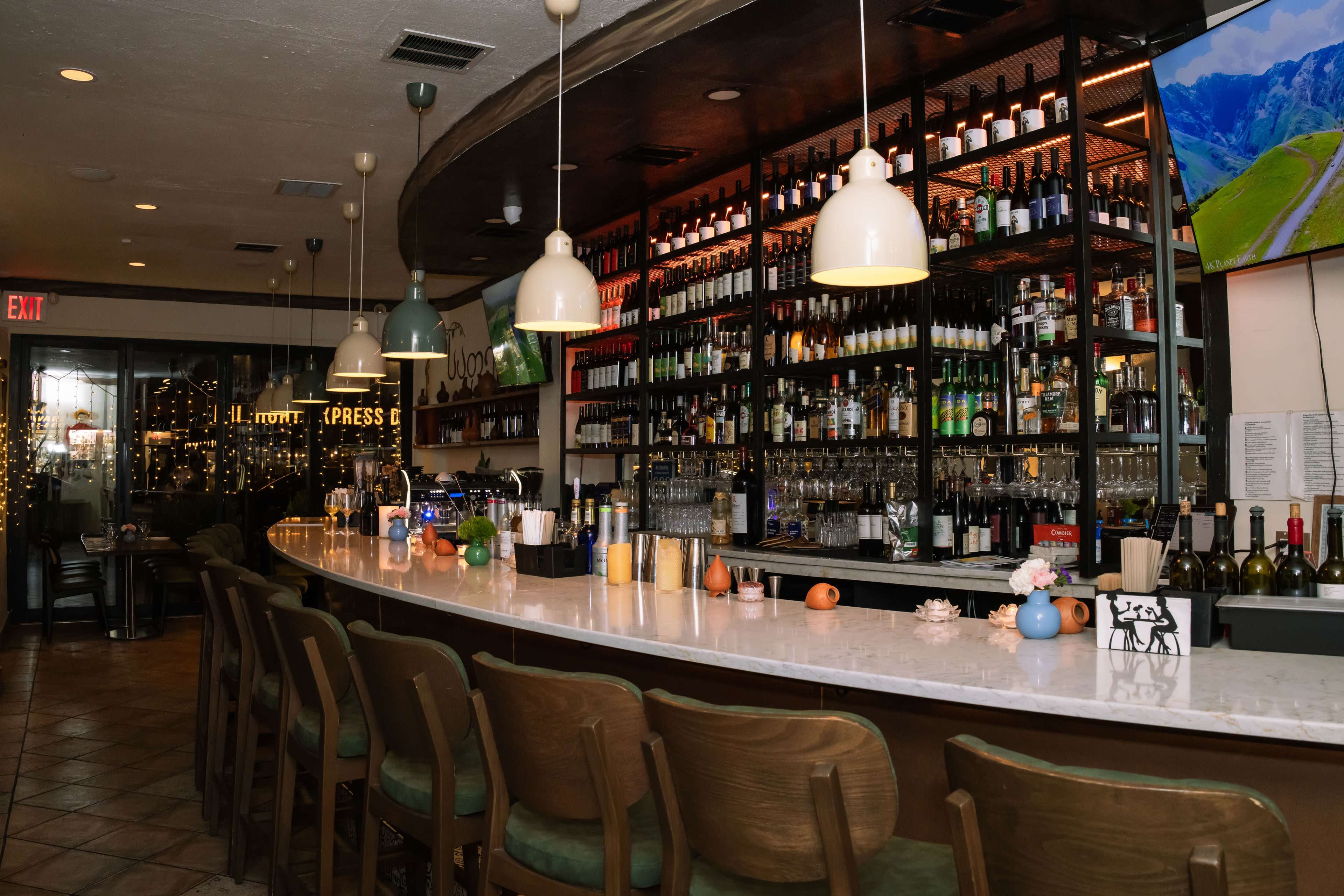 The image shows a well-lit bar area with a long marble counter and a large selection of bottles displayed on shelves behind the bar.