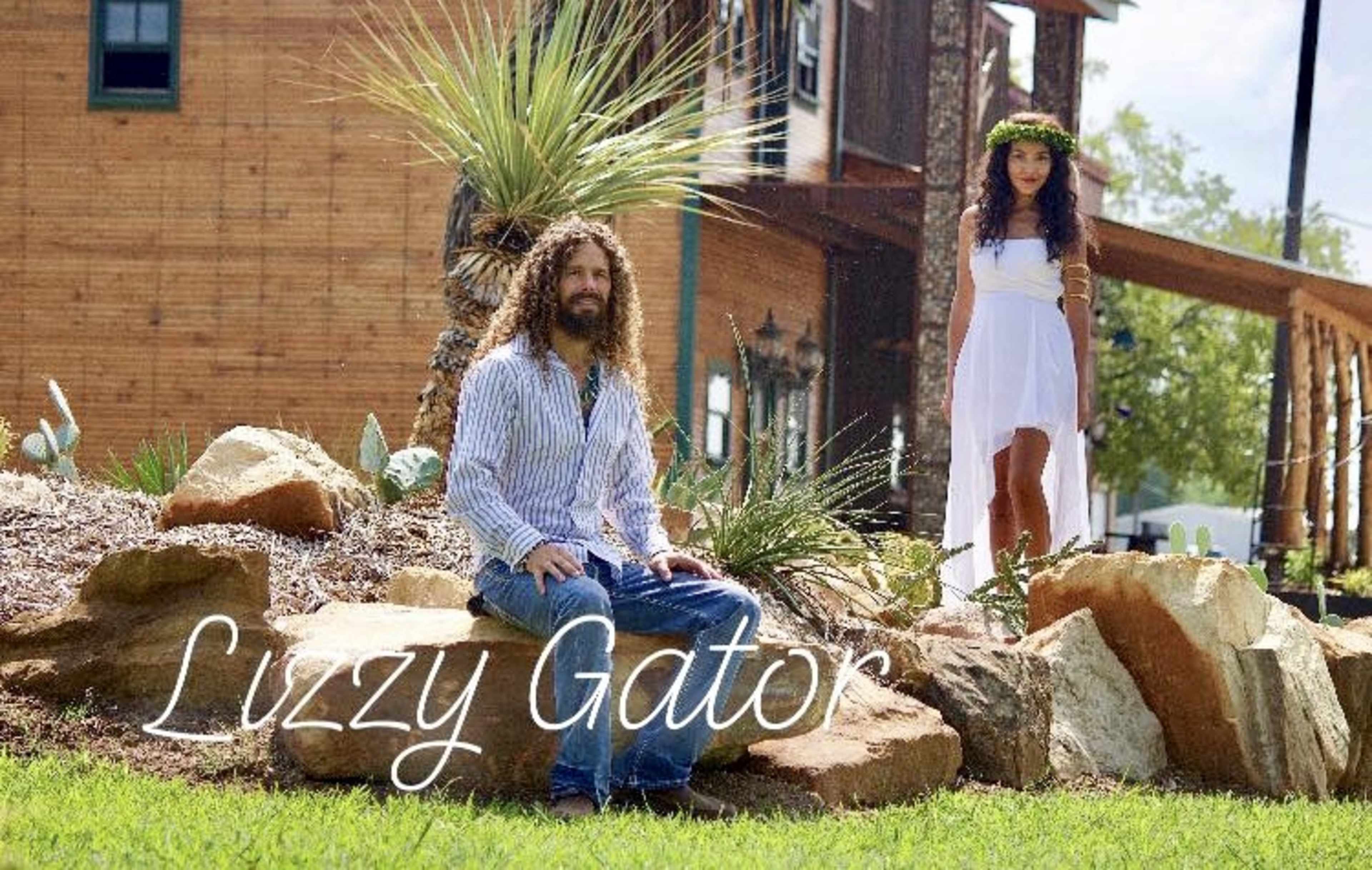 A man with long hair sits on a rock while a woman in a white dress stands nearby in a landscaped yard with plants and a wooden building in the background.