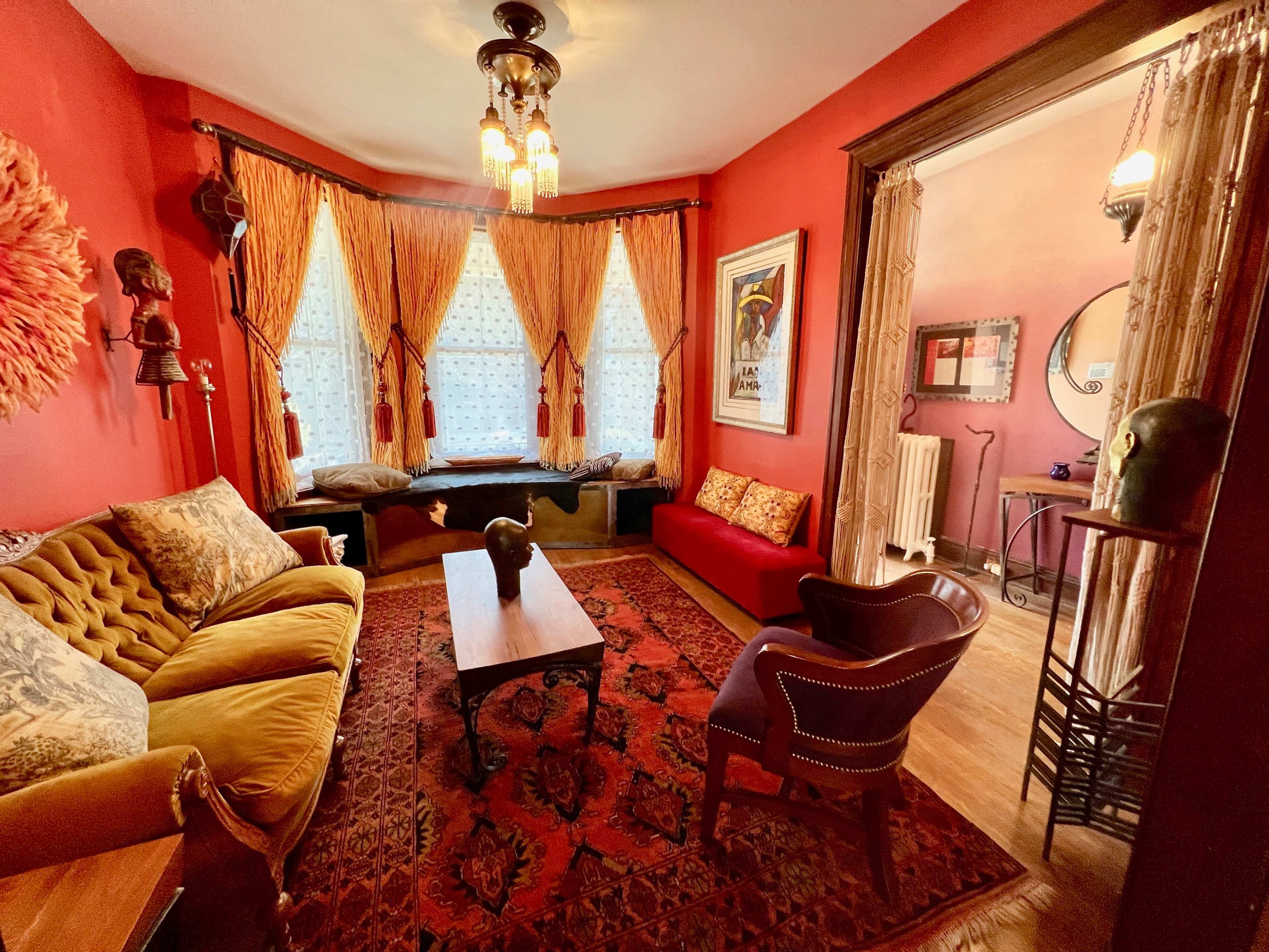 A cozy living room with red walls, a brown patterned rug, a vintage sofa, and large windows dressed with ornate curtains.