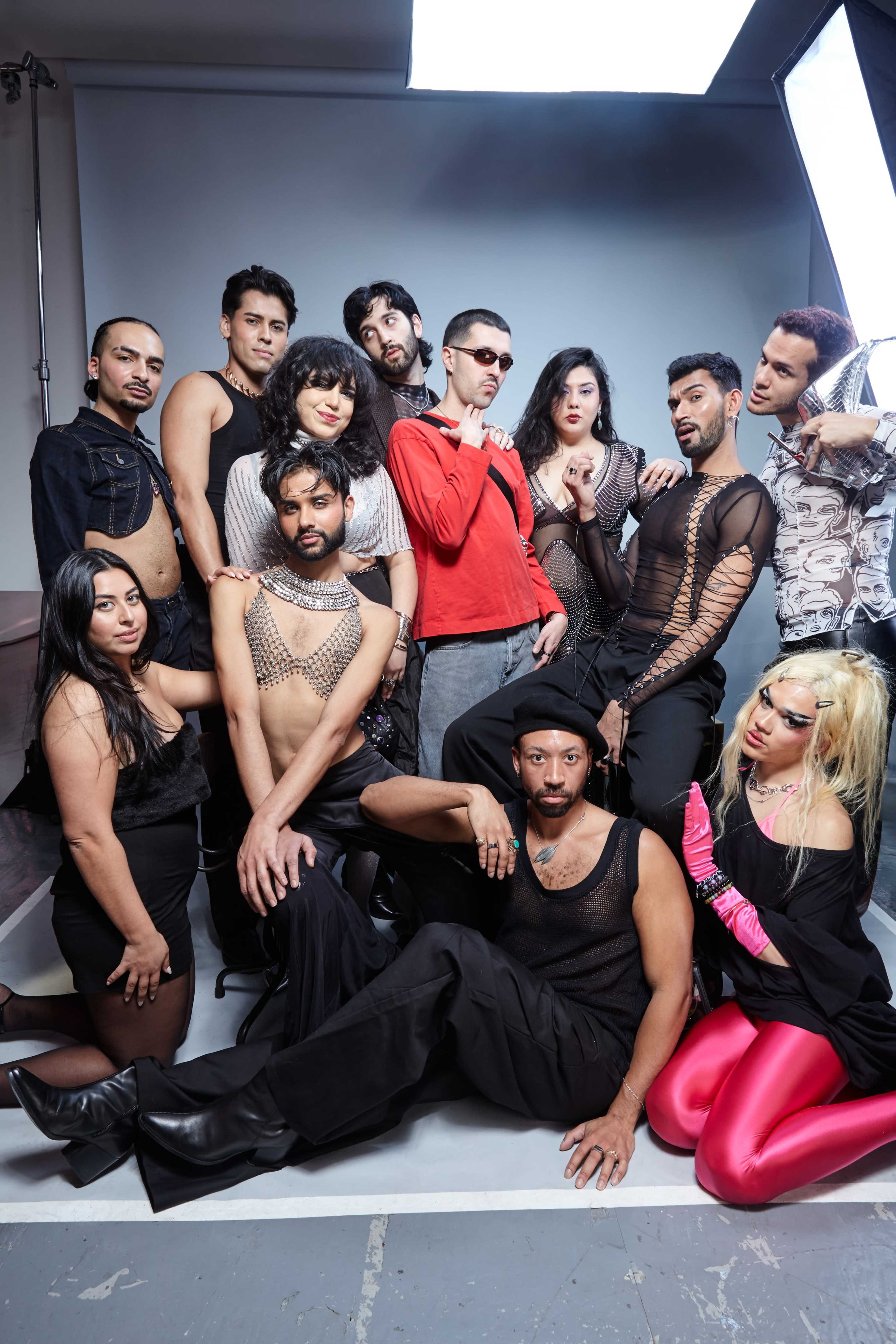 A diverse group of twelve people poses together in a studio setting, with a mix of bold fashion choices and expressions.