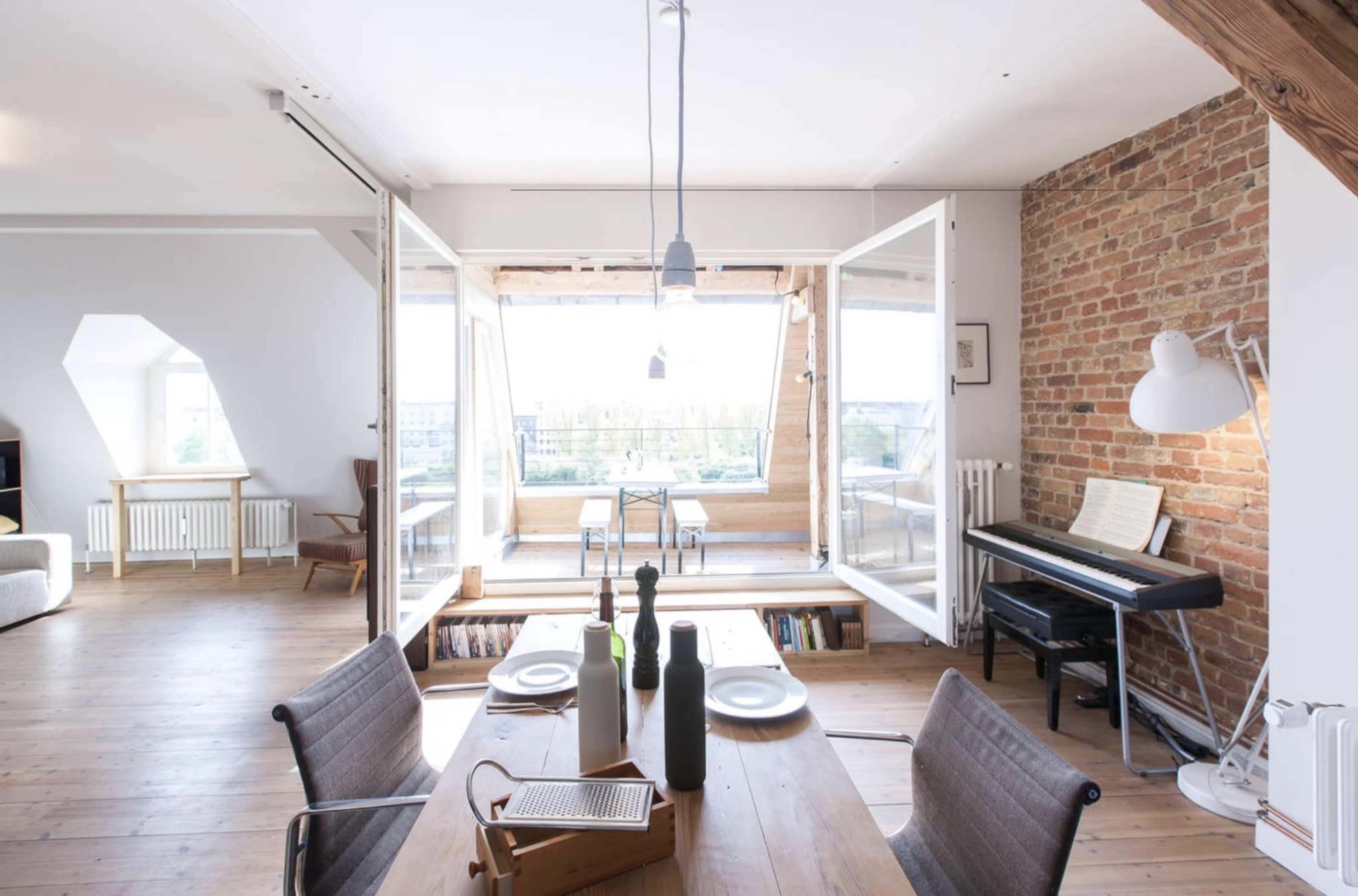 A dining area features a wooden table set with plates, facing a bright room with large windows that open to a balcony, while exposed brick walls and wooden beams add character to the space.