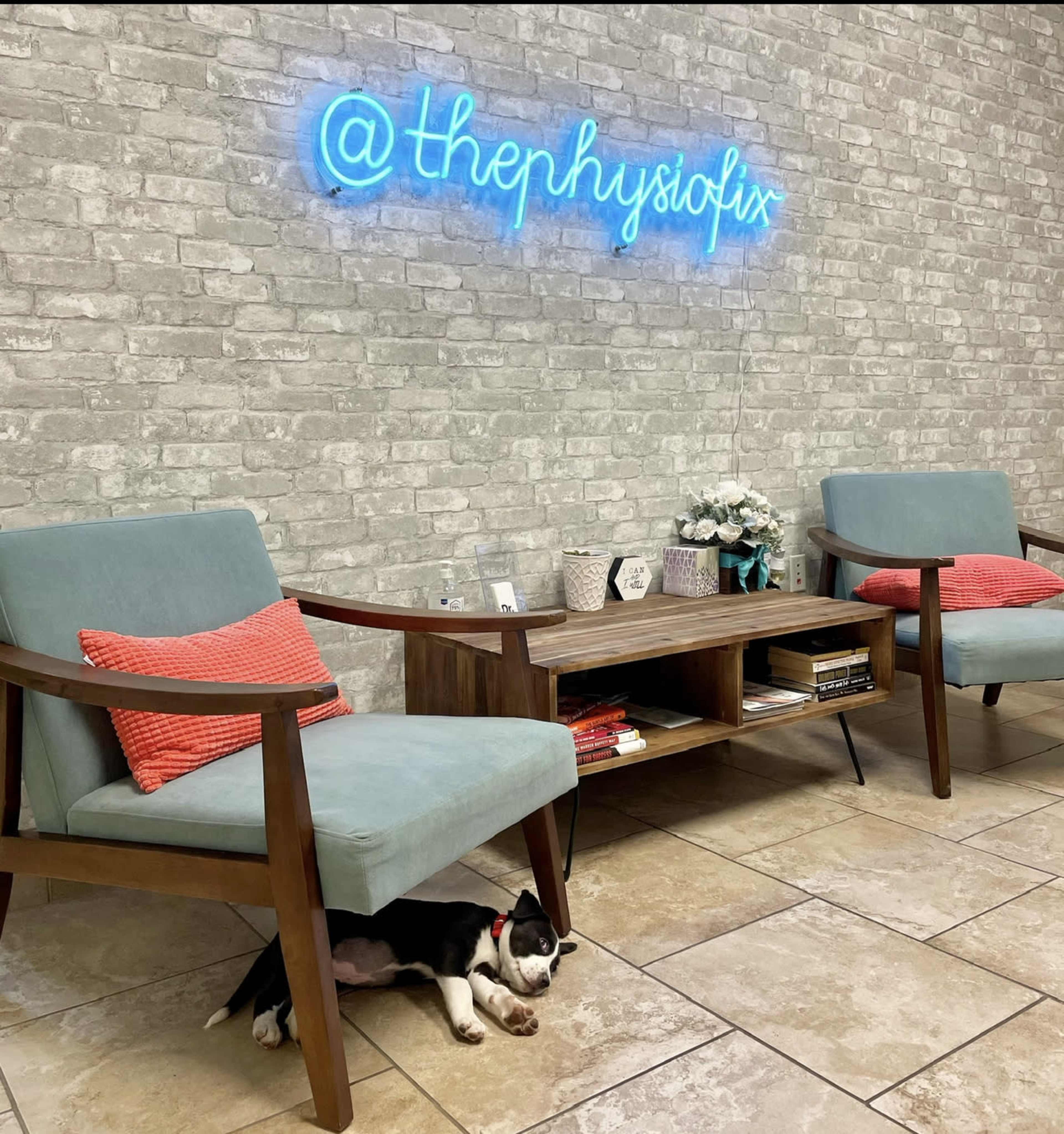 A small dog rests under a wooden table in a waiting area with two blue chairs and a neon sign on a brick wall.